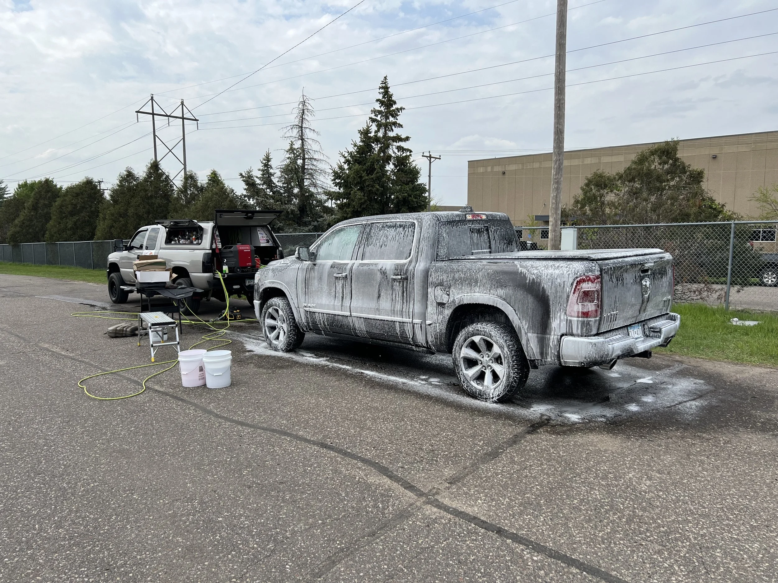 A black pickup truck is being washed with foam on its exterior in an outdoor setting, with another silver pickup truck nearby and various cleaning supplies and equipment around.