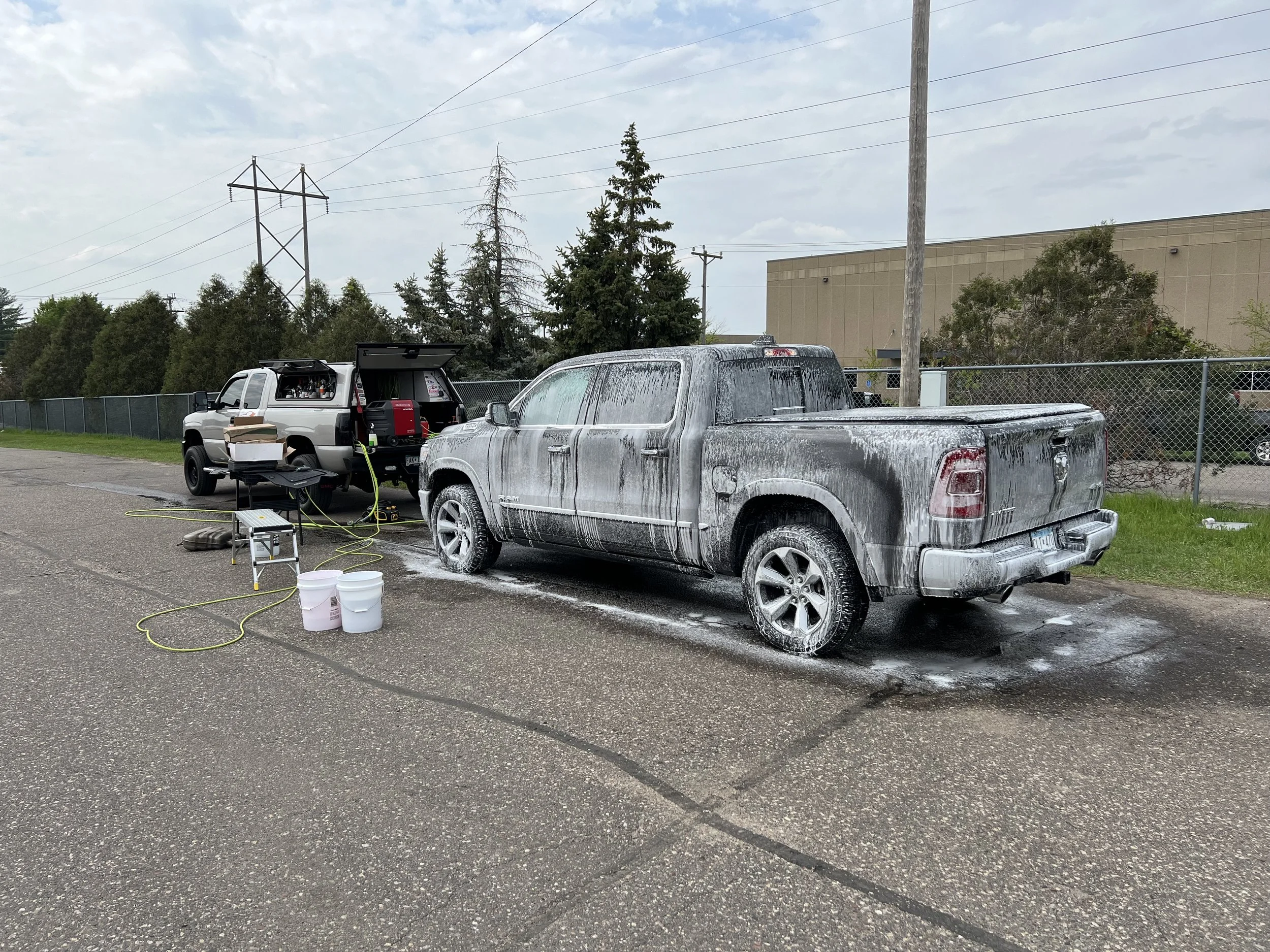 A pickup truck being washed, covered in soap suds, parked on a wet parking lot with a car wash setup beside it.