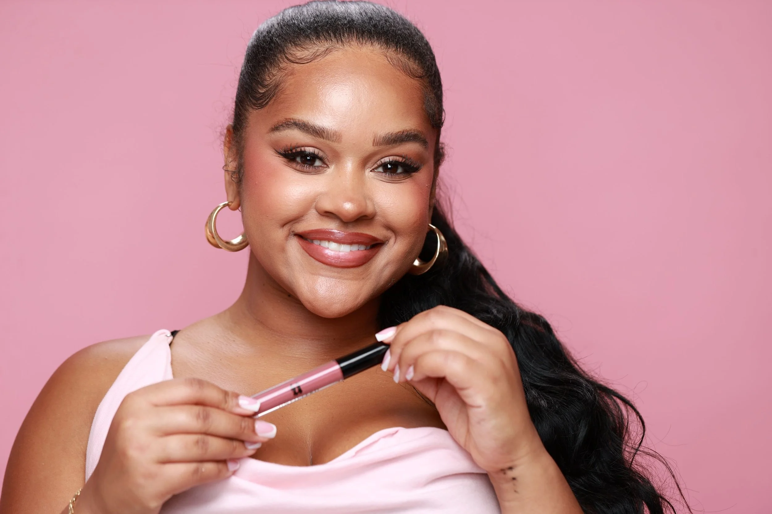 A young woman with dark hair and hoop earrings smiling while holding a pink lip gloss tube against a pink background.