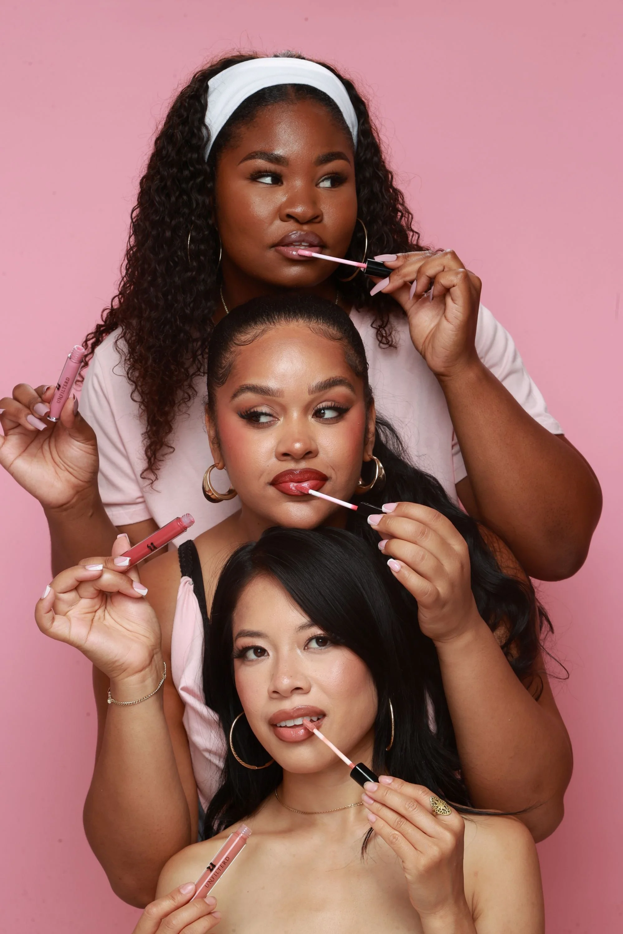 Four diverse women applying lip gloss together against a pink background, with each holding a lip gloss tube.
