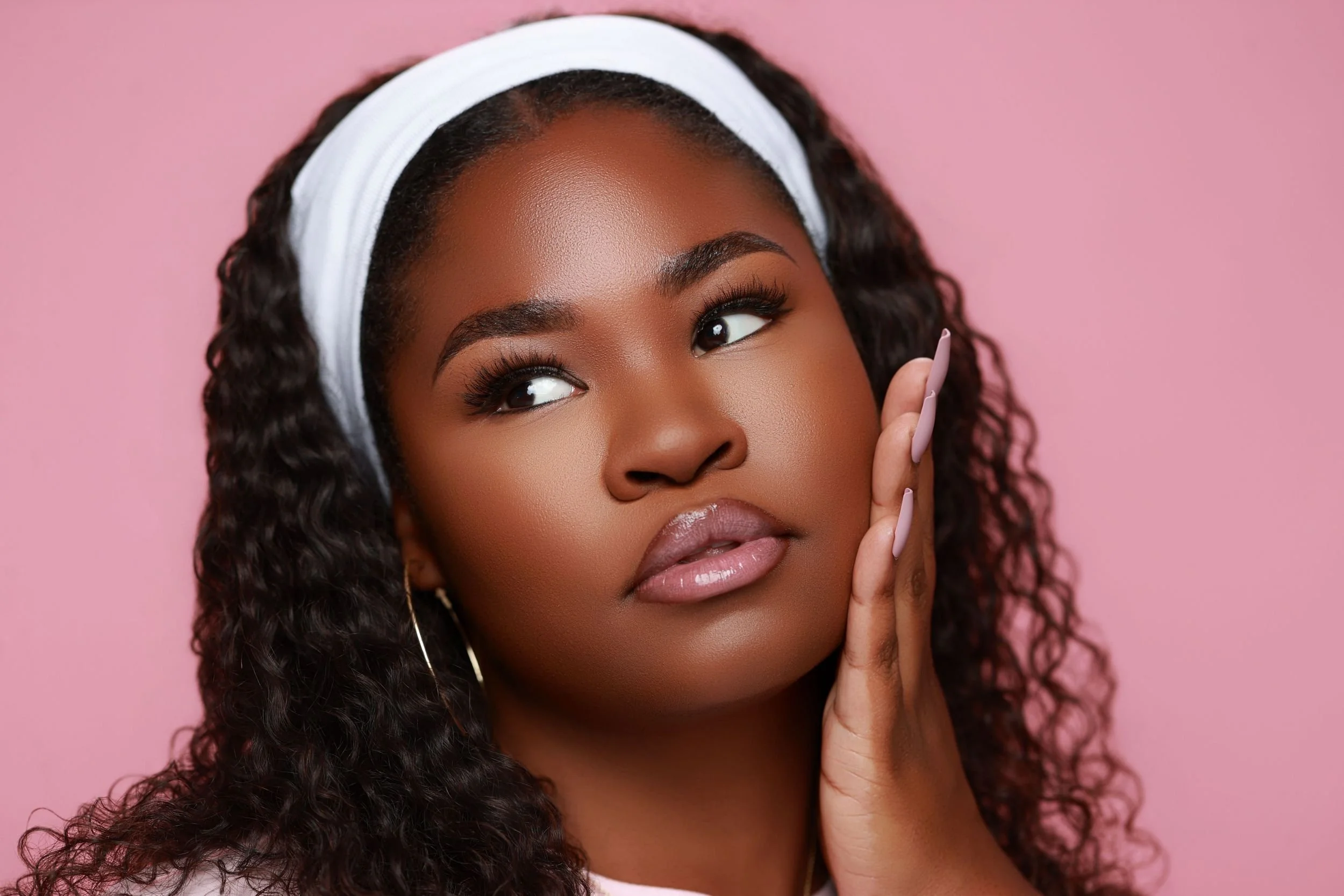 Close-up of a woman with dark, curly hair wearing a white headband and makeup, touching her face, against a pink background.