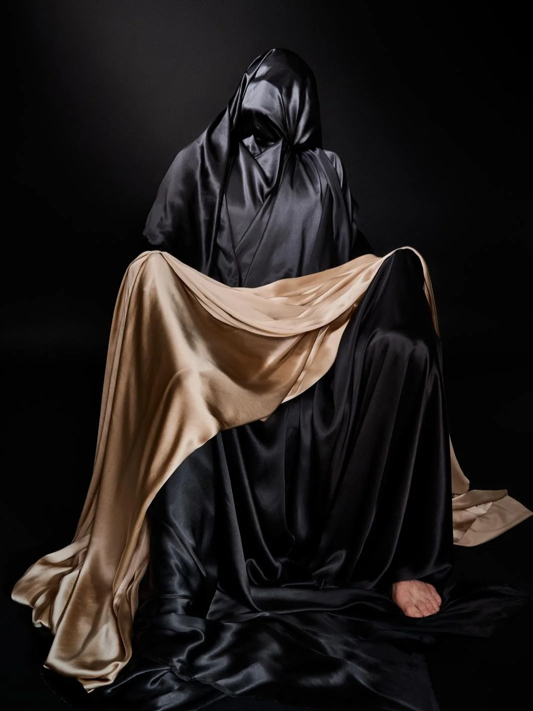 Two figures covered in black and beige satin fabric, one seated and the other with a hood, against a black background.