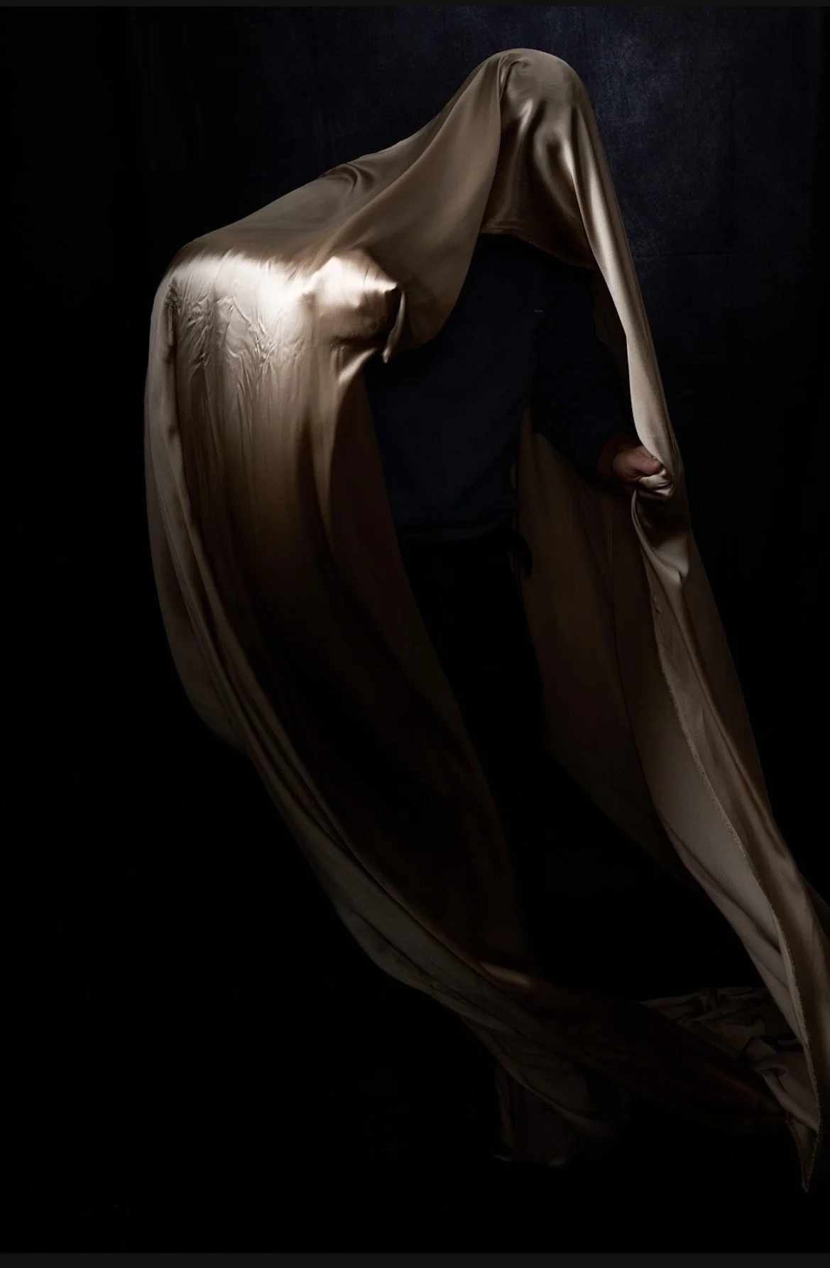 Person standing behind a long, flowing satin sheet that drapes over their head and body, with dark background.