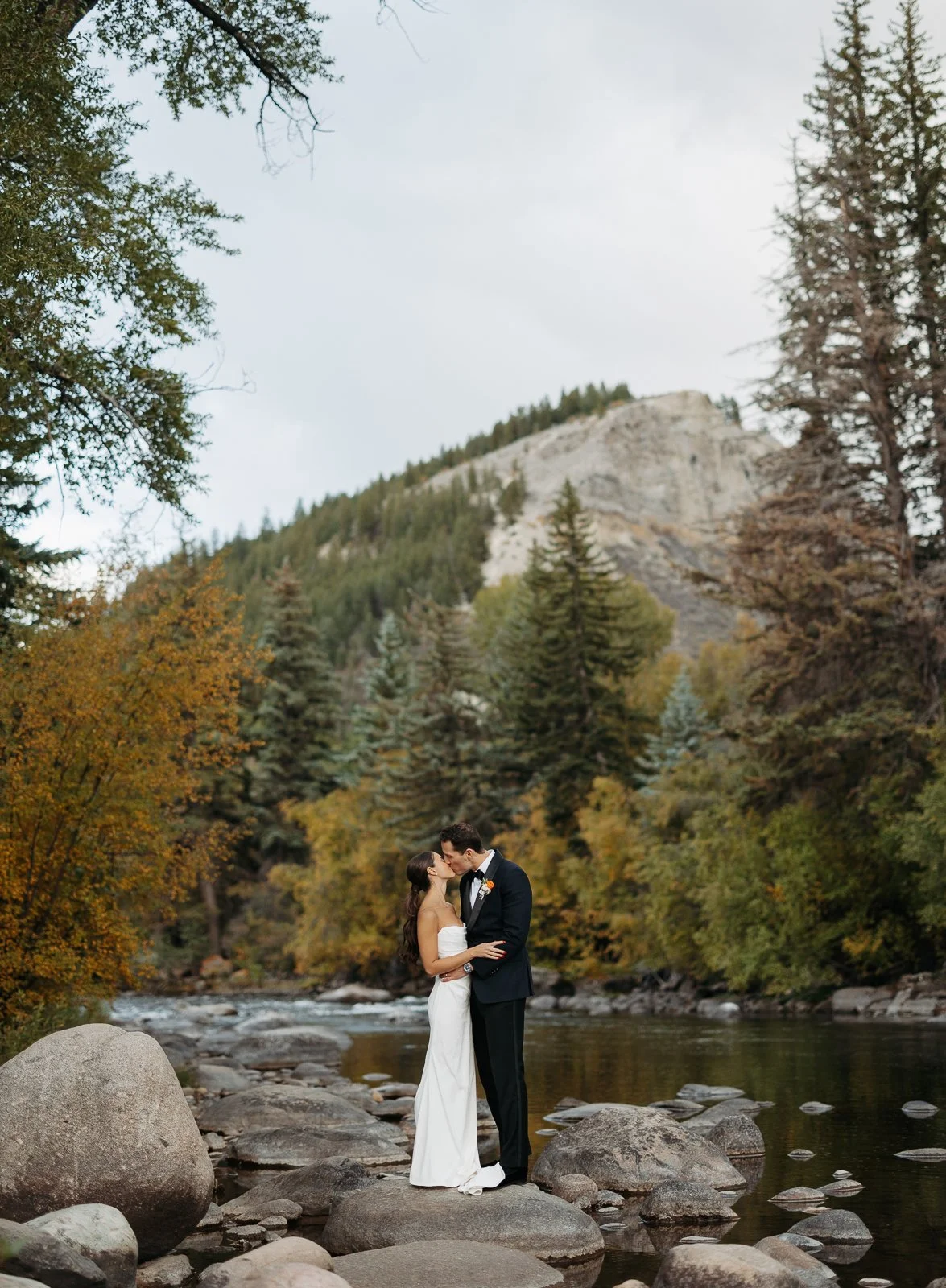 A bride and groom stand on rocks in a river, kissing, surrounded by trees with fall foliage and a mountain in the background.
