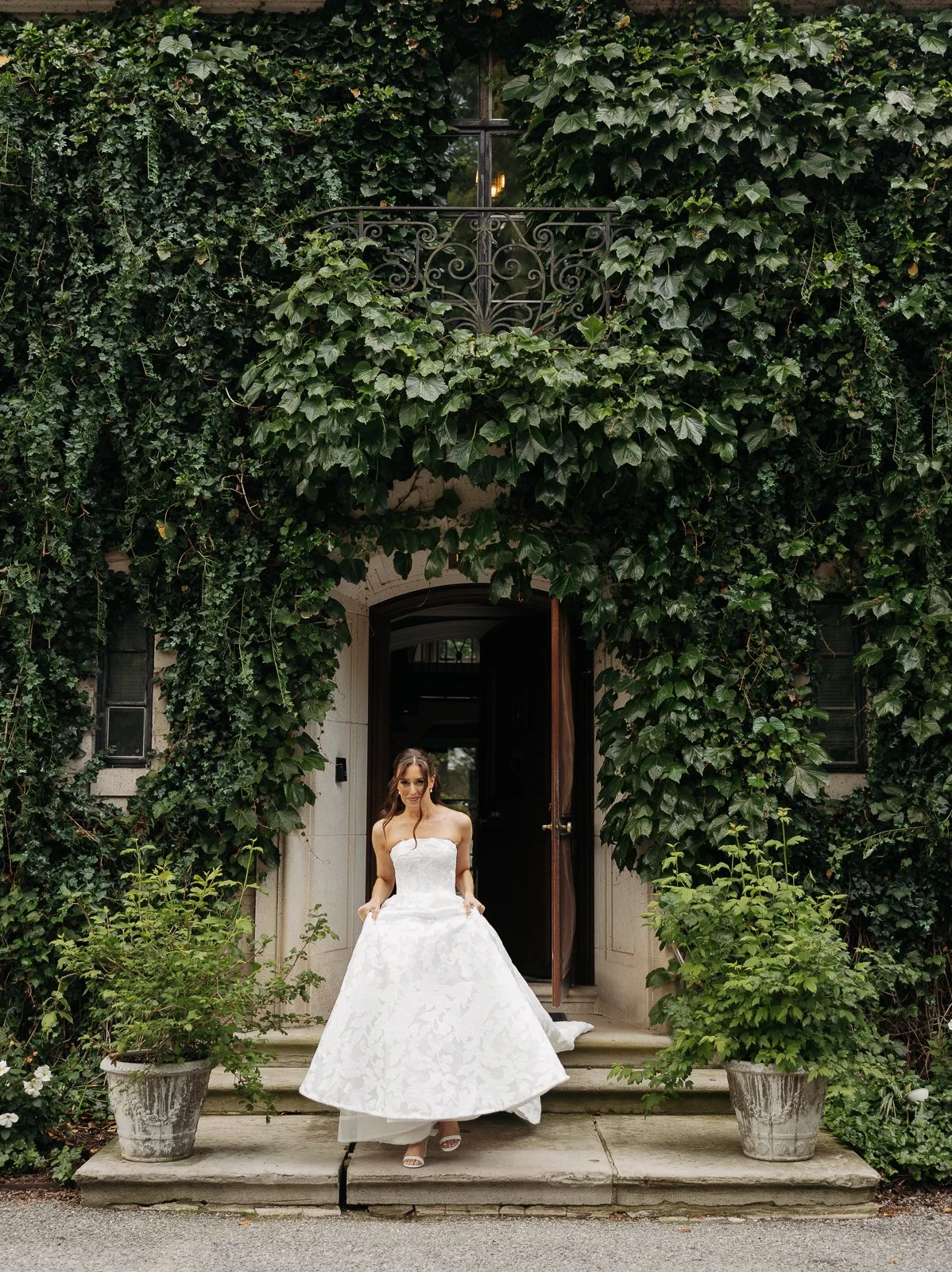 A woman in a strapless white wedding gown standing on porch steps in front of a door, surrounded by lush green ivy and potted plants.