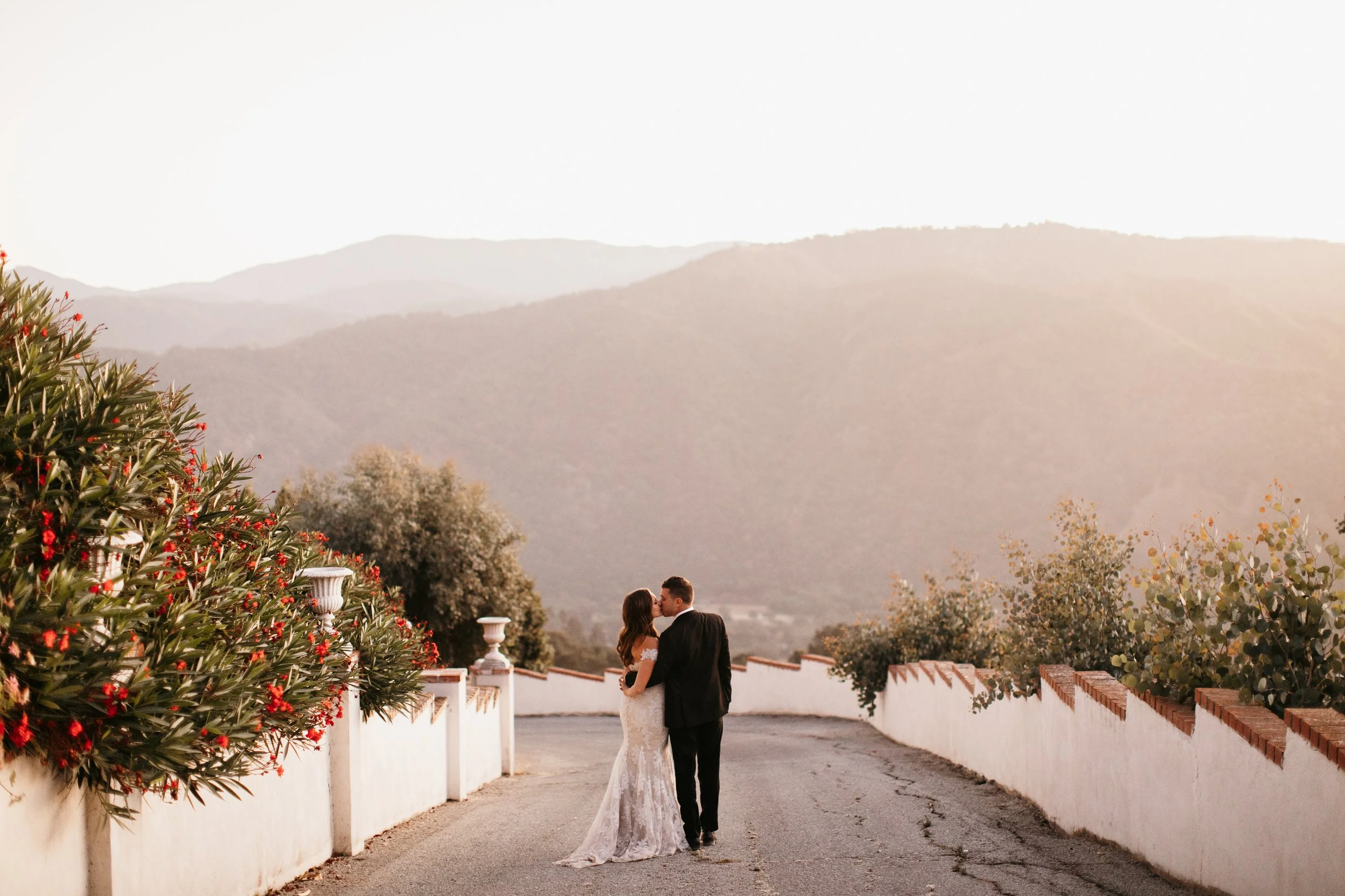 A bride and groom kiss outdoors on a paved path, with a mountain range in the background and greenery on the sides. The bride wears a white wedding gown and the groom is in a dark suit.