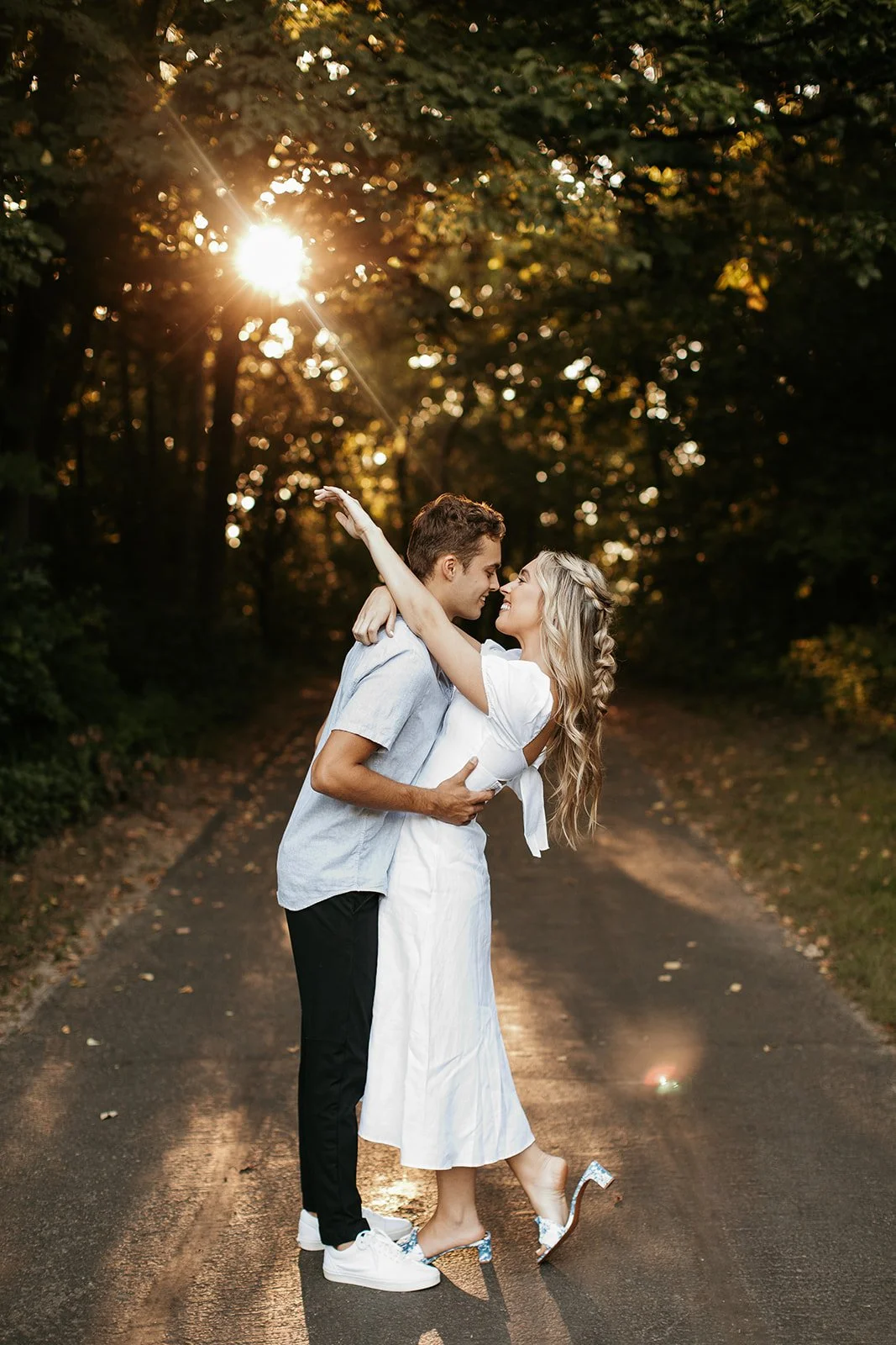 A couple embraces and smiles closely on a rural road surrounded by trees at sunset.
