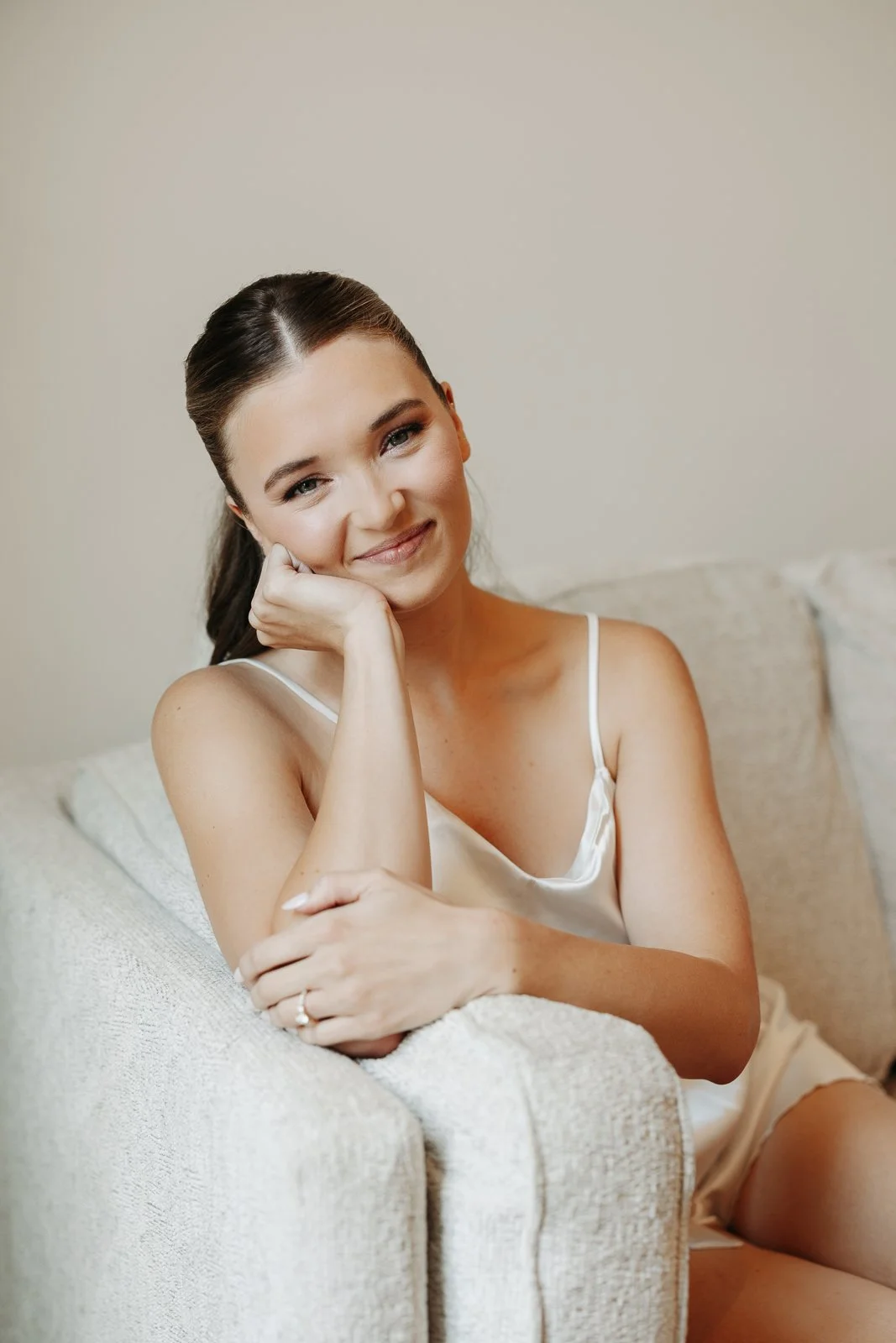 A woman with brown hair in a ponytail sitting on a beige sofa, smiling softly, wearing a satin camisole, resting her head on her hand.