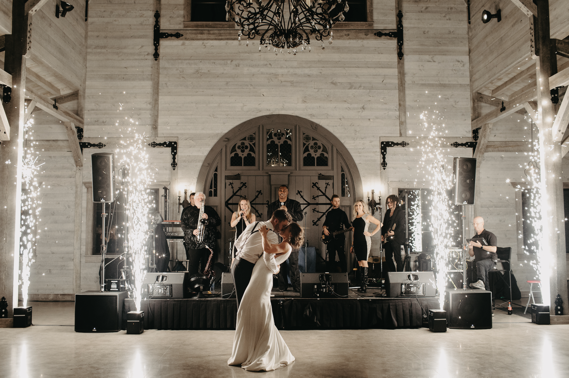 Bride and groom dancing at a wedding reception with live band on stage, sparklers on sides, chandelier hanging from ceiling, in a rustic venue with light wood walls and arched window.