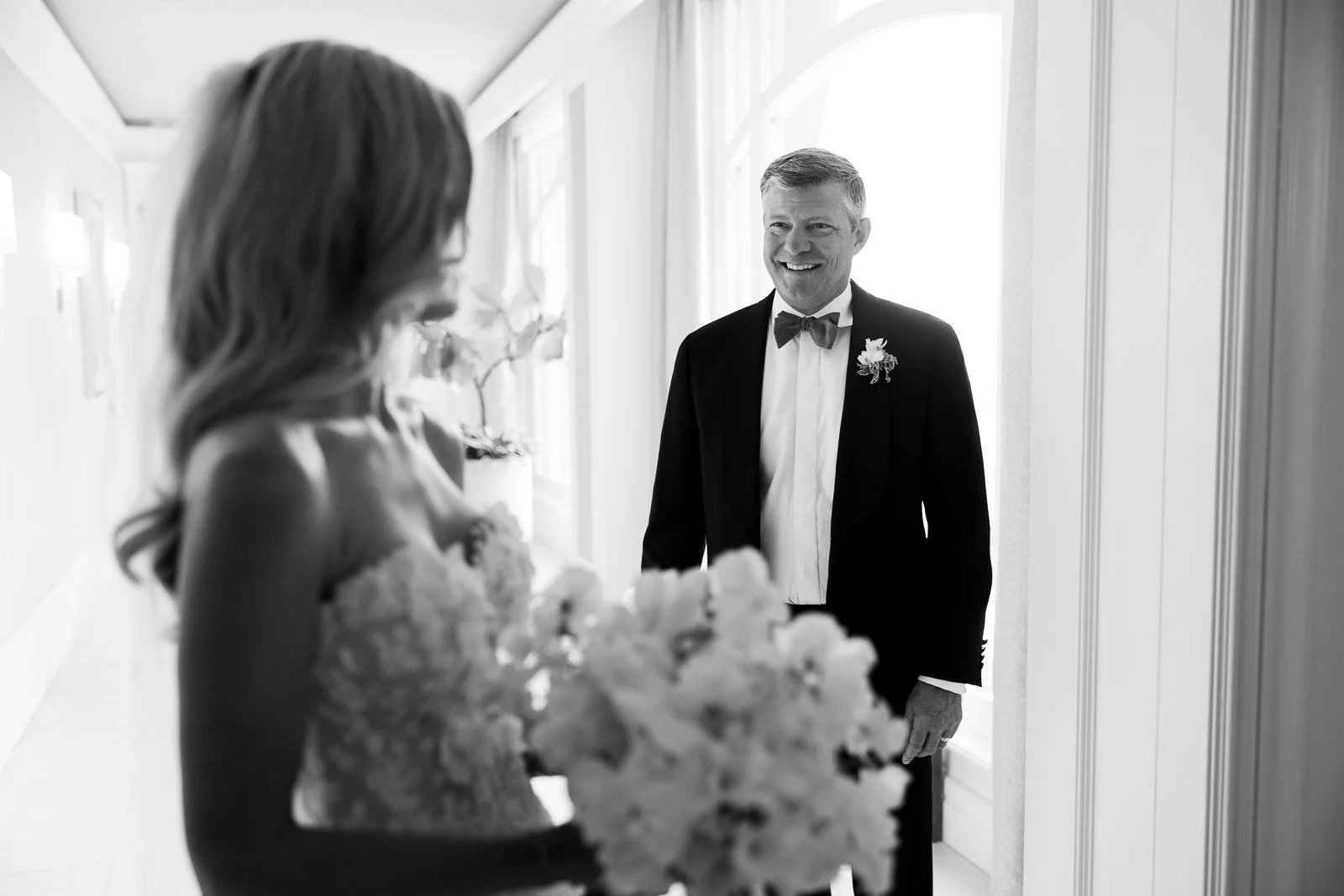 A groom in a tuxedo with a bow tie smiling at a bride holding a bouquet of flowers in a brightly lit hallway.