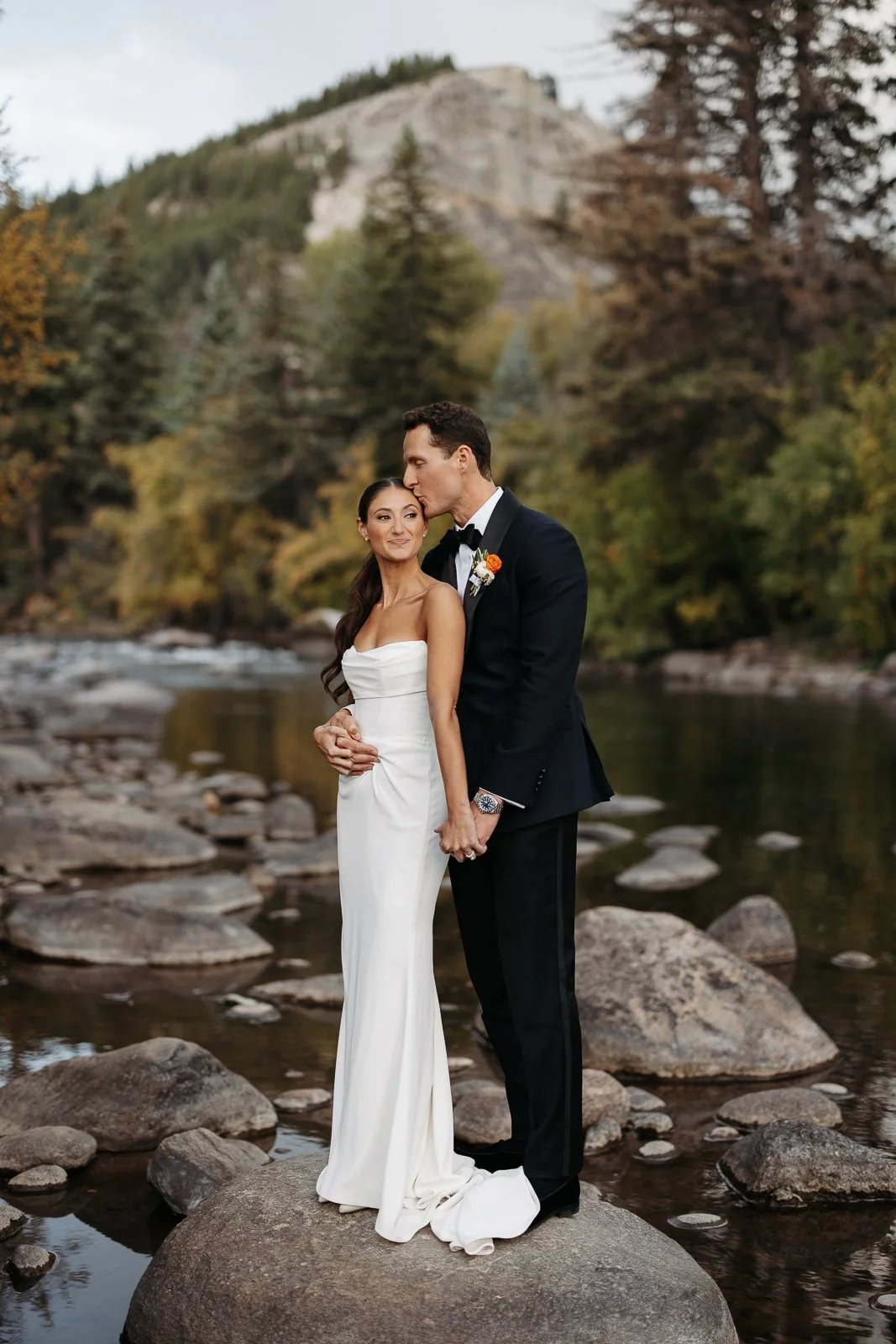 A newlywed couple standing on a large rock in a river, surrounded by trees and mountains, with the groom kissing the bride on the head.