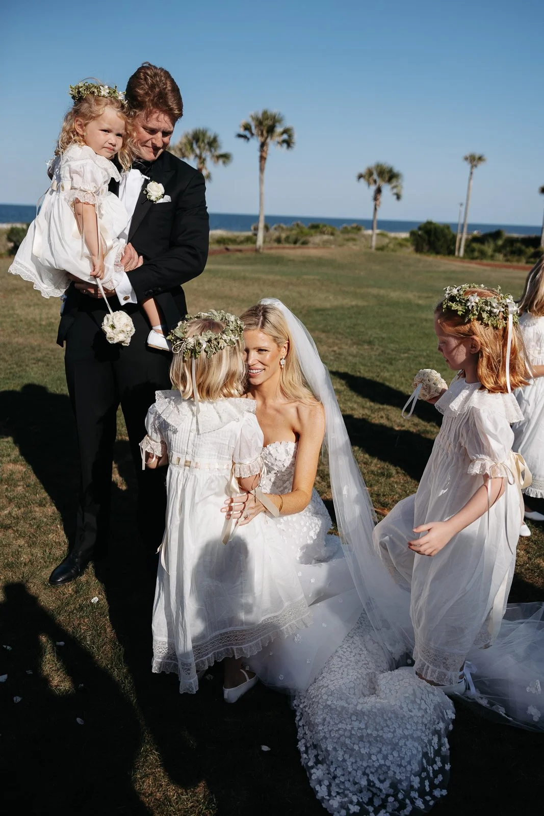 A wedding ceremony outdoors near the beach with women in white dresses and floral crowns, a man in a tuxedo holding a young girl, and a woman in a wedding gown kneeling and smiling at a child.