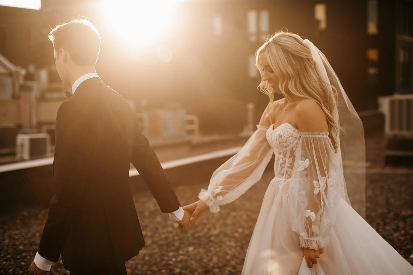 A bride and groom holding hands and walking together outdoors at sunset, with the bride wearing a white wedding dress and the groom in a black tuxedo.