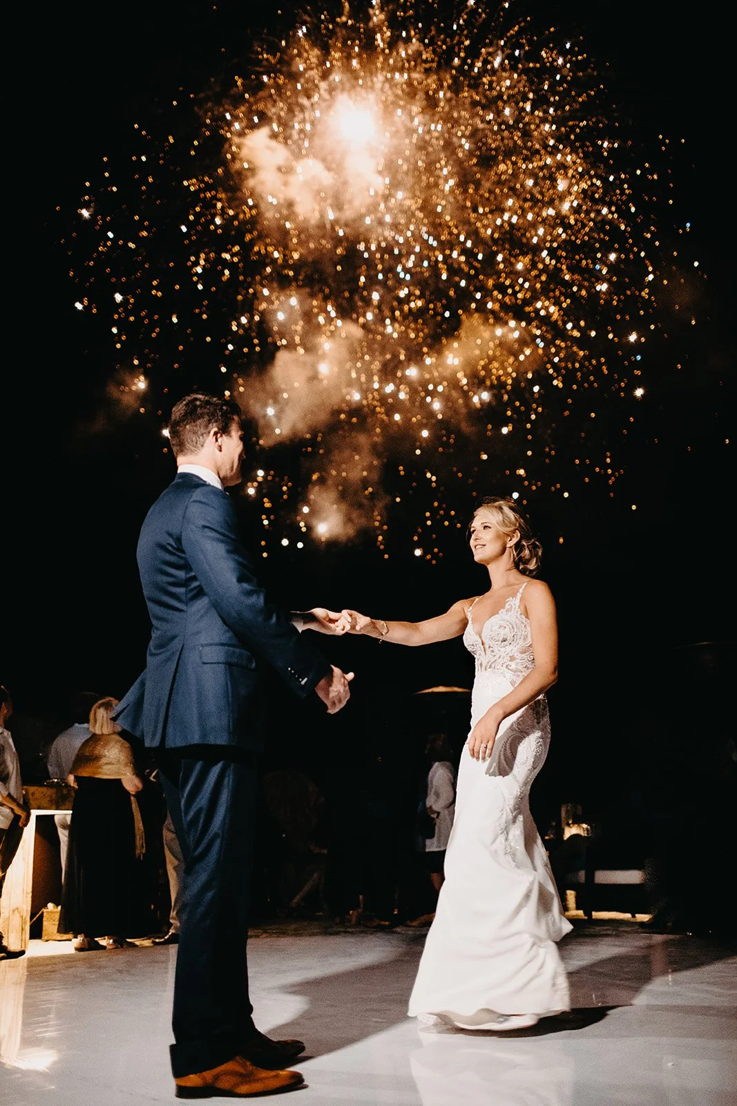 A bride and groom holding hands during their wedding dance under a sky with fireworks lighting up the night.