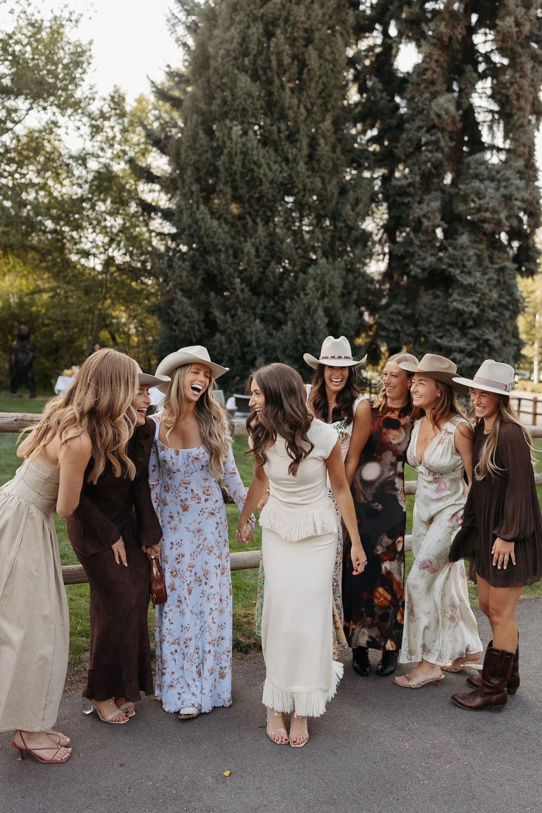 Group of women wearing dresses and hats, laughing and talking outdoors near a wooden fence with trees in the background.