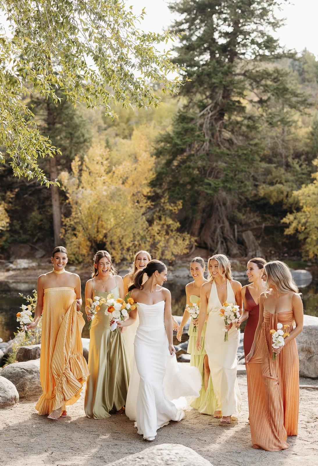 Group of women in colorful dresses holding bouquets, walking and smiling outdoors near a river with trees and rocks.