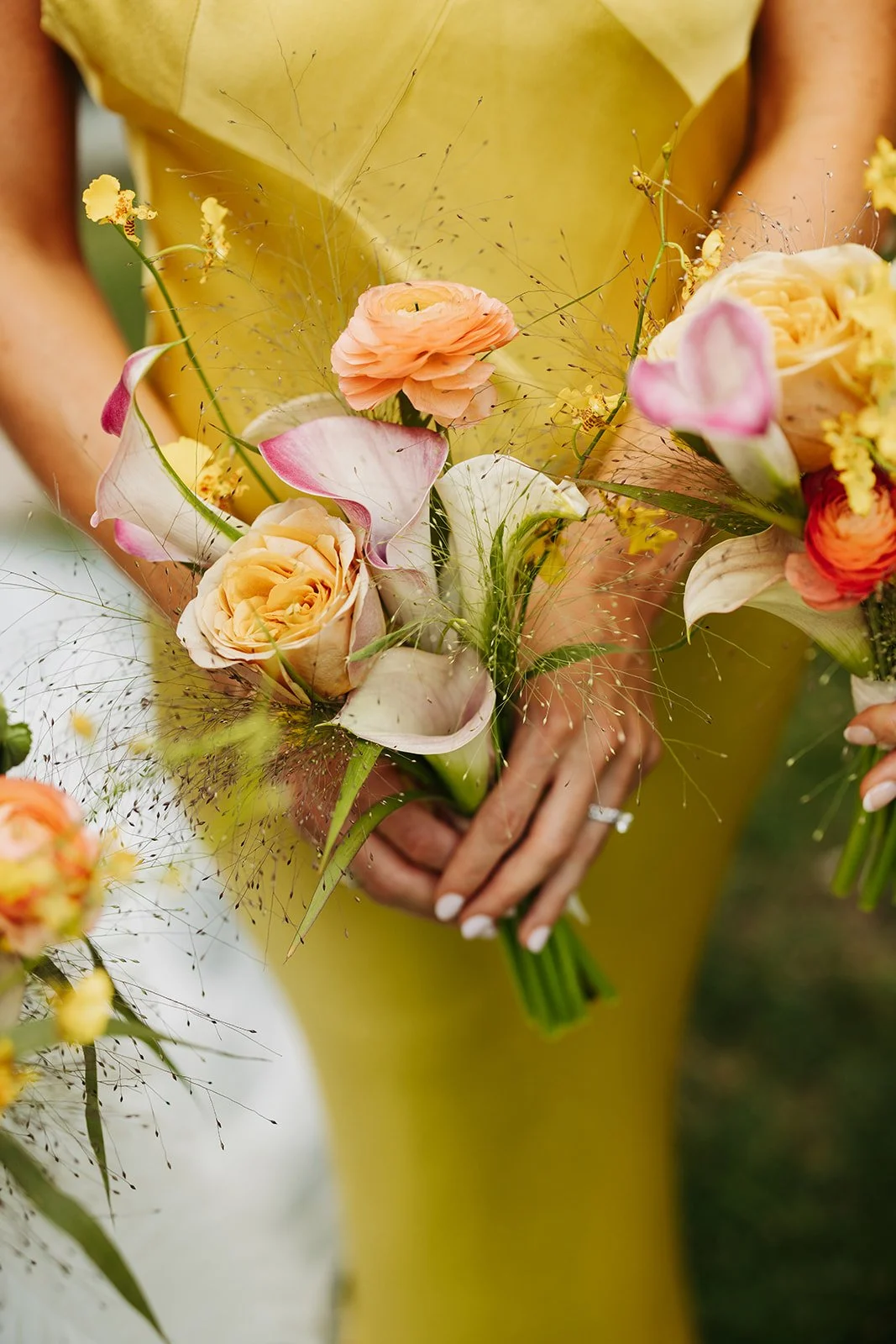 Close-up of a person holding a bouquet of pastel-colored flowers, including roses and calla lilies, with greenery and delicate sprigs.