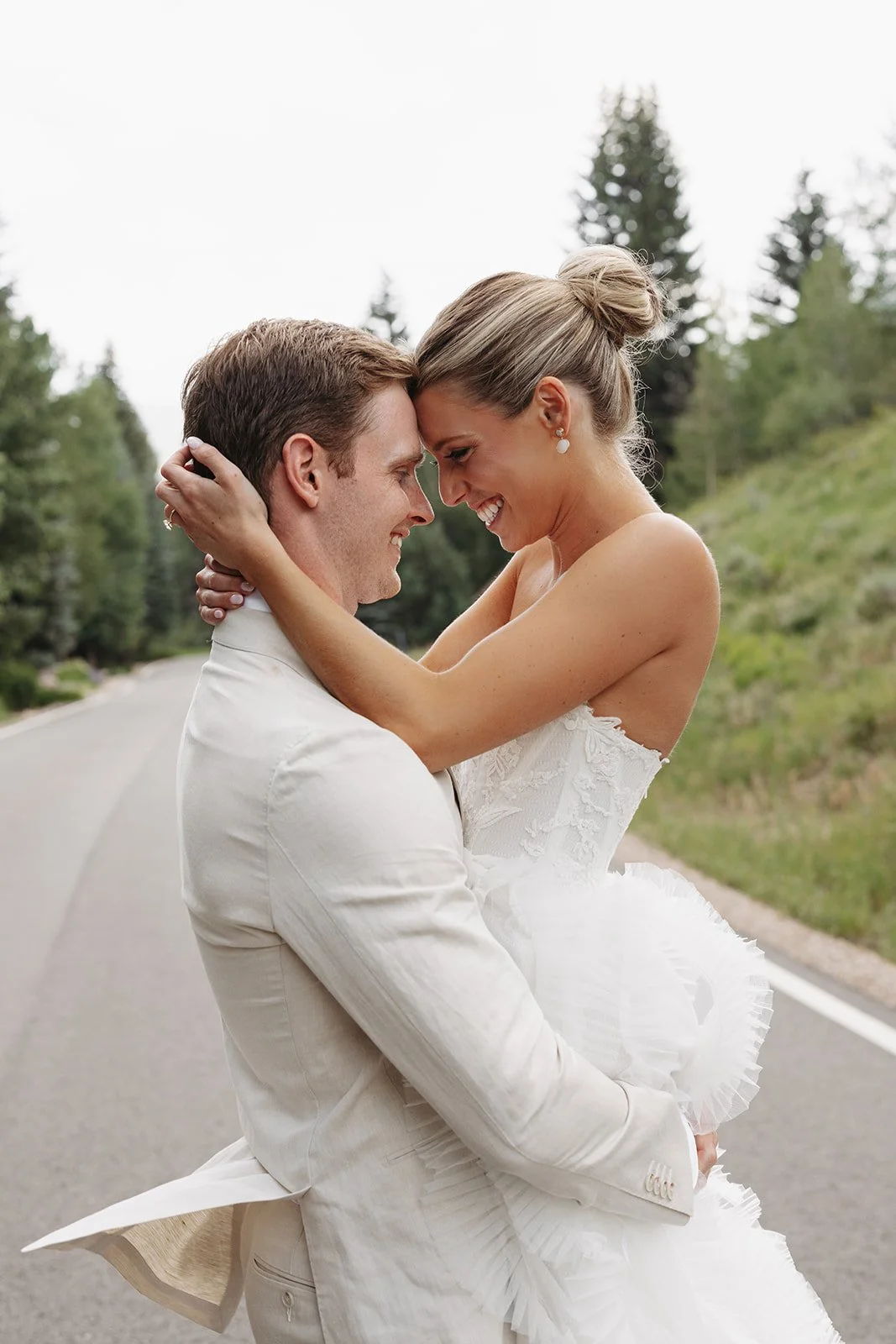 A couple dressed in wedding attire embracing on a road with trees in the background.