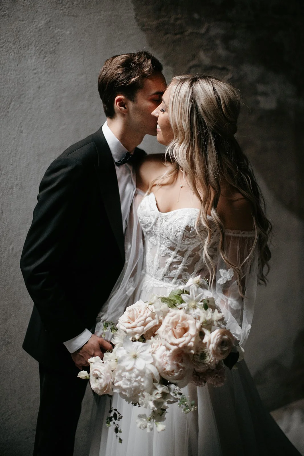 A bride and groom are kissing, with the groom in a black suit and the bride in a white wedding dress, holding a large bouquet of pink and white flowers.