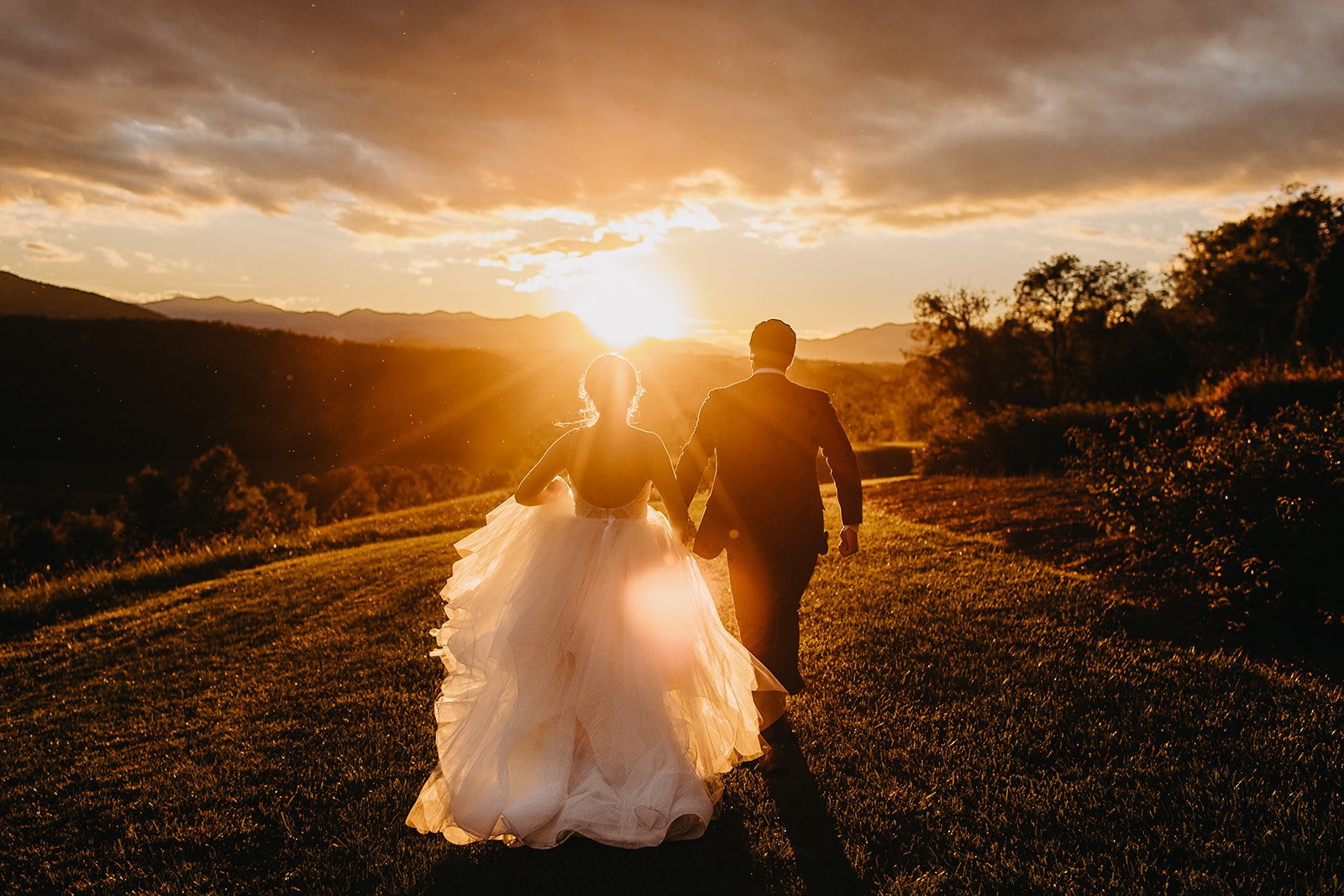 A bride and groom walking hand in hand outdoors during sunset, with the bride in a white wedding gown and the groom in a suit, scenic landscape in the background.