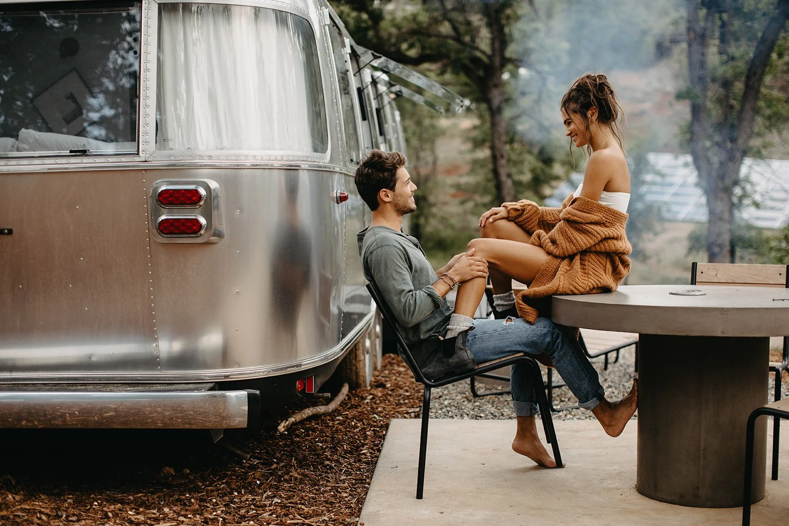 A young man and woman sit together outside near an Airstream trailer, smiling and enjoying each other's company, surrounded by trees in a natural setting.