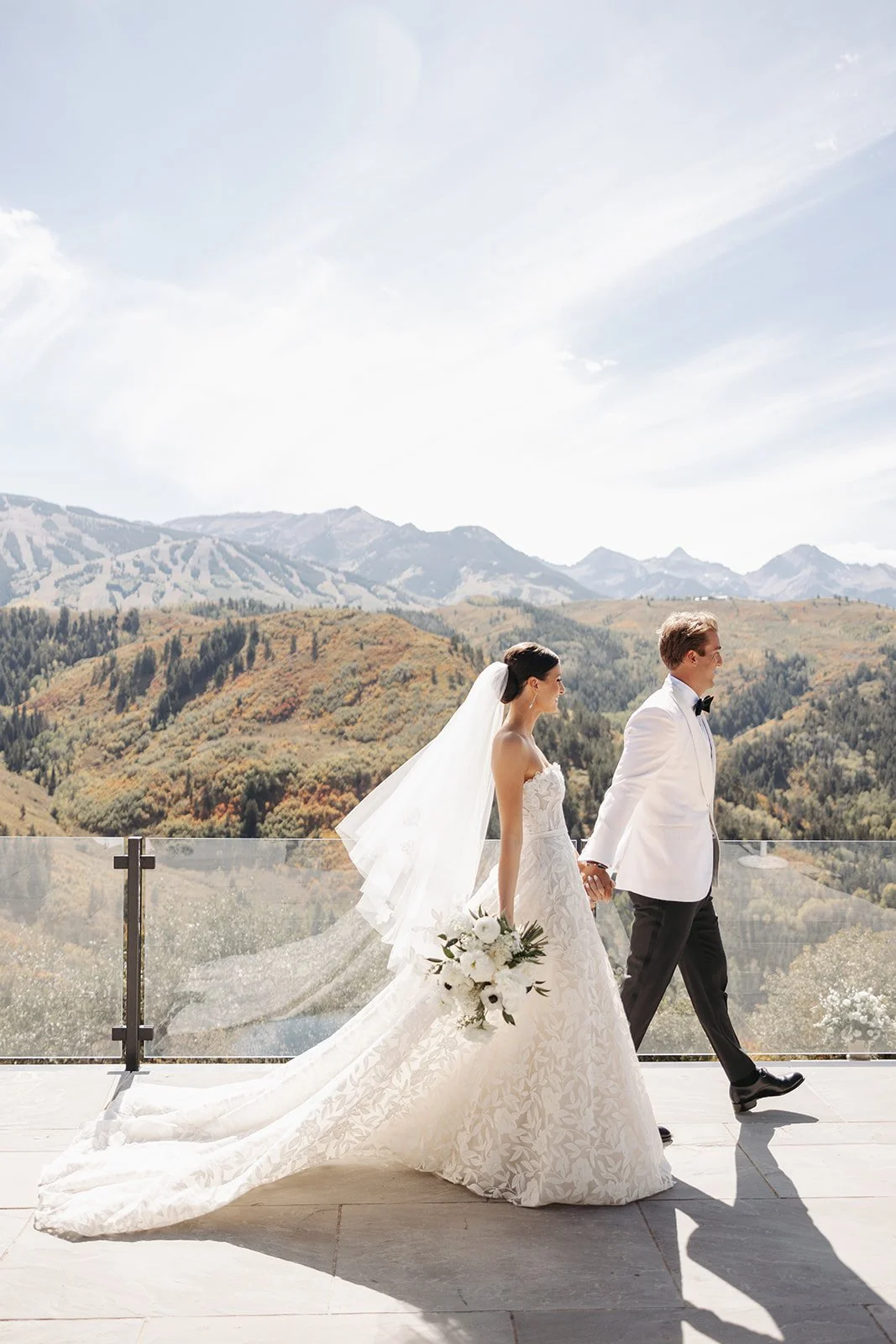 A bride and groom walking hand in hand outdoors on a balcony with mountain scenery in the background, the bride in a white wedding gown holding a bouquet, and the groom in a white tuxedo with a black bow tie.