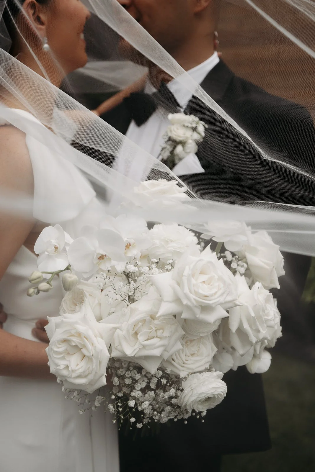 A bride and groom on their wedding day, partially visible, with the bride holding a bouquet of white roses and other white flowers, both dressed in formal wedding attire.