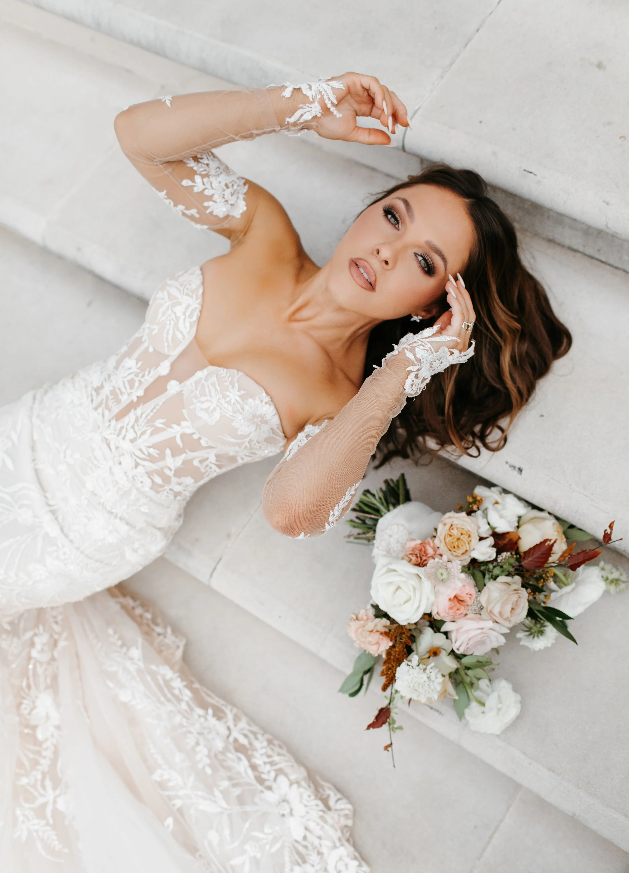 A woman in a white lace wedding dress lying on steps with a bouquet of white, peach, and blush flowers beside her.