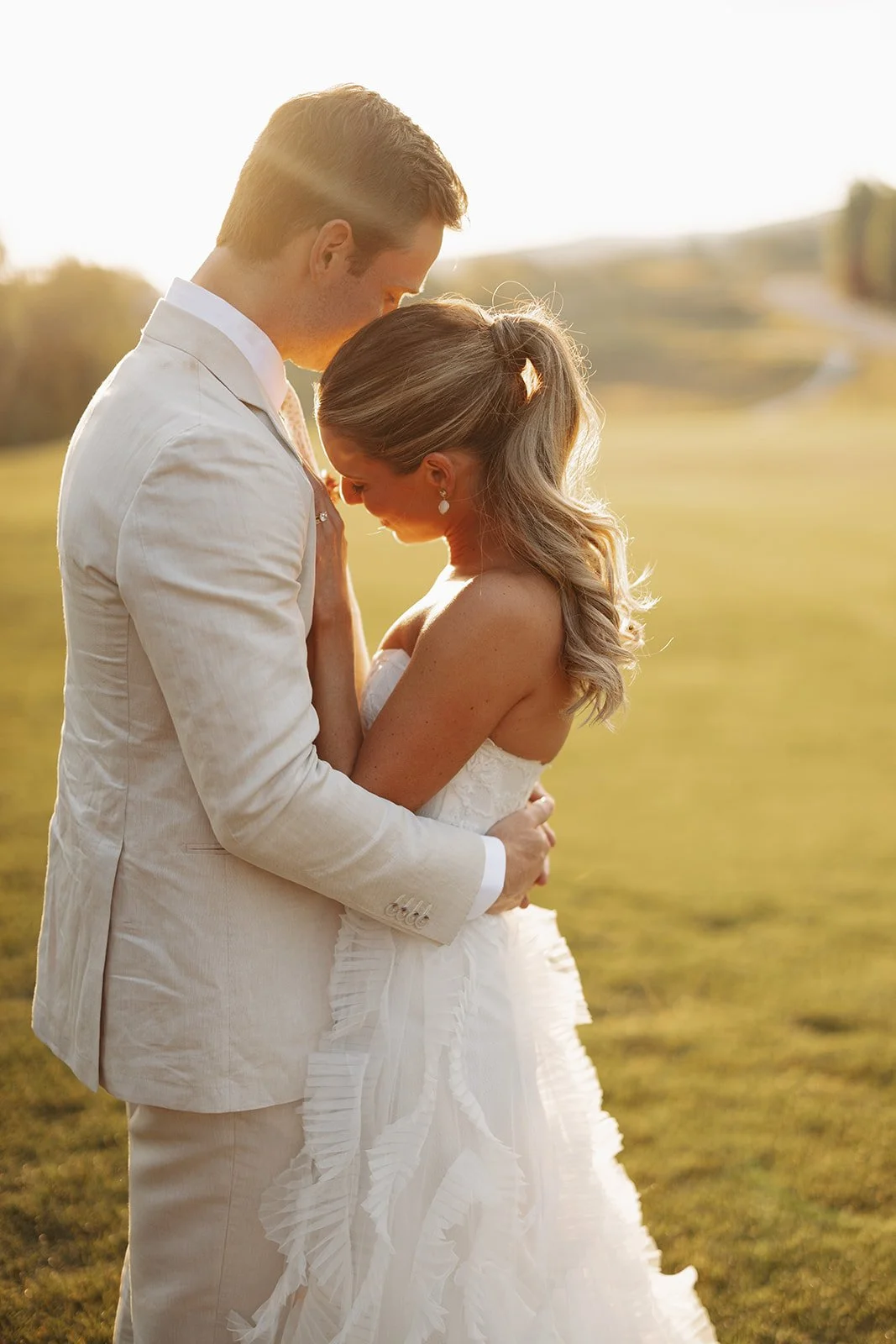 A couple dressed in wedding attire embracing outdoors during sunset, with a scenic landscape in the background.