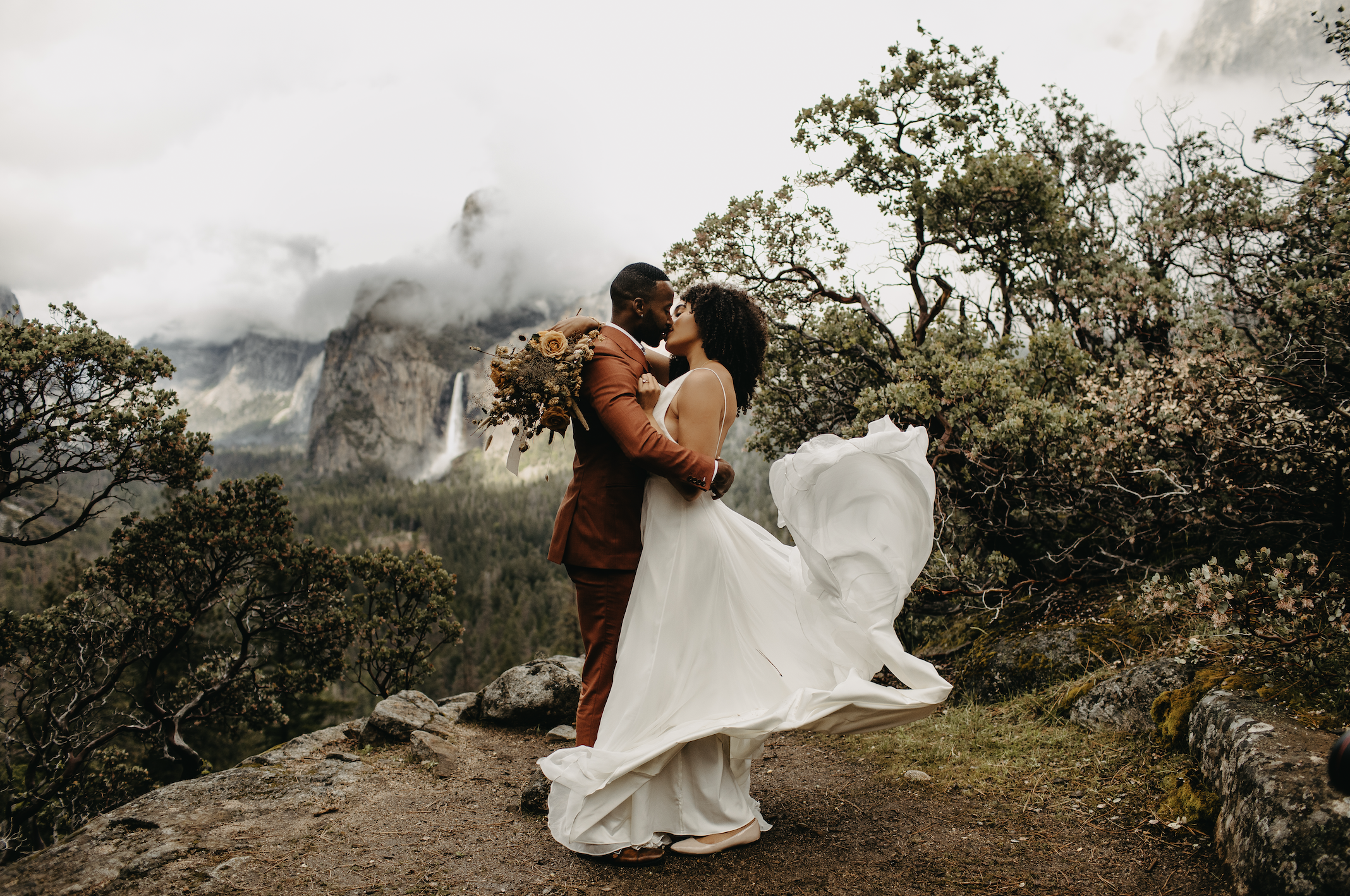 A couple dressed in wedding attire sharing a kiss outdoors amidst trees and rocky terrain, with mountains and cloudy sky in the background.