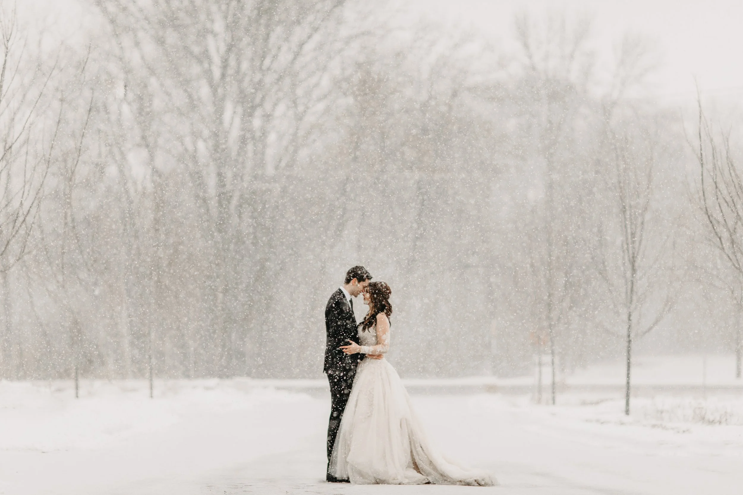A couple dressed in wedding attire stands close together in a snowy outdoor setting, surrounded by snow-covered ground and bare trees, with snow falling around them.