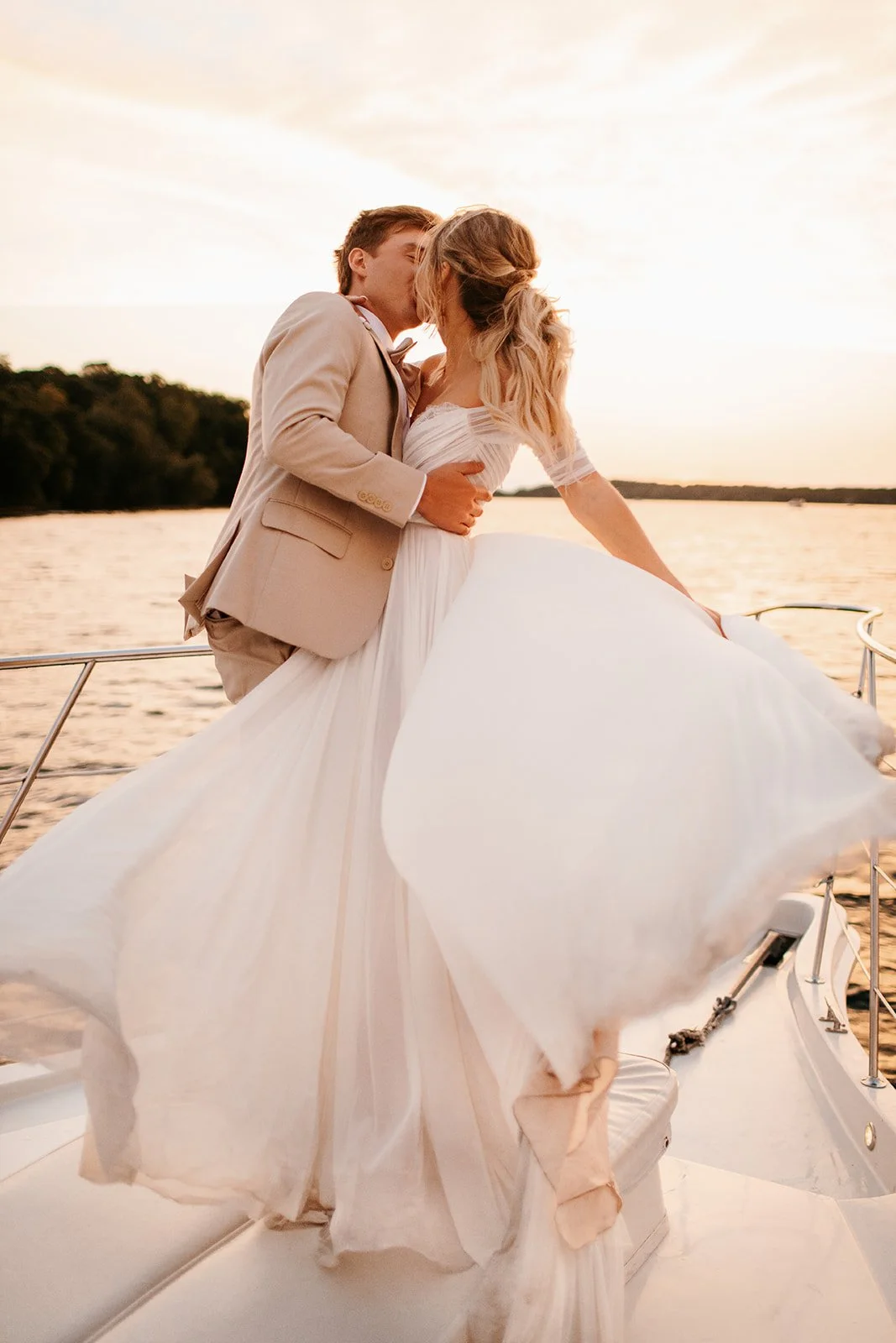 A bride and groom sharing a kiss on a boat at sunset with water and trees in the background.