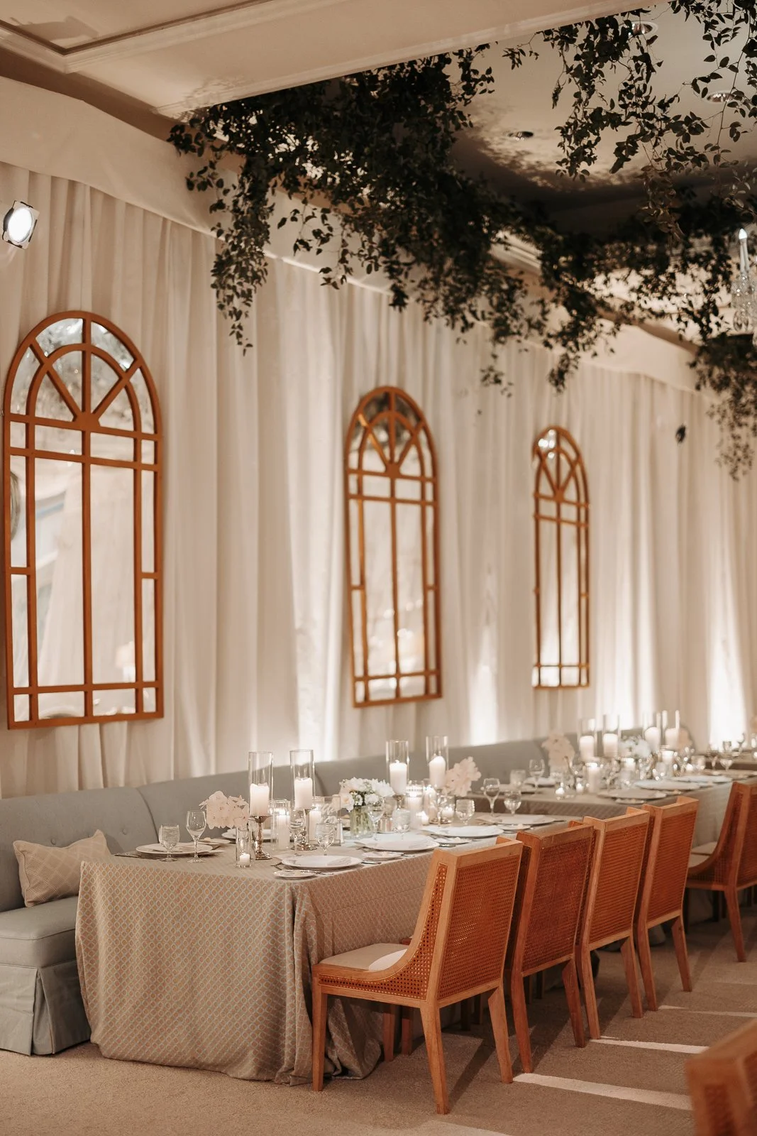 Elegant banquet table setup with candles, white flowers, glassware, and china in a decorated room with white curtains, arched windows, and hanging greenery.