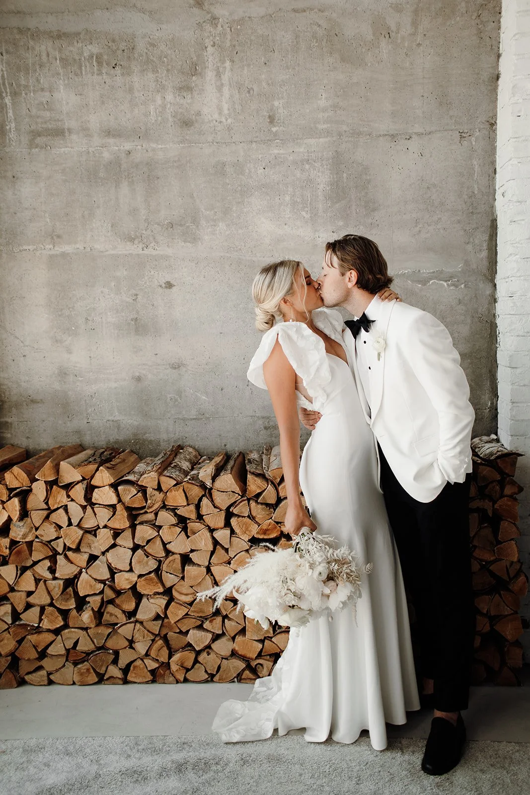 A bride and groom kissing in front of a concrete wall with stacked firewood.