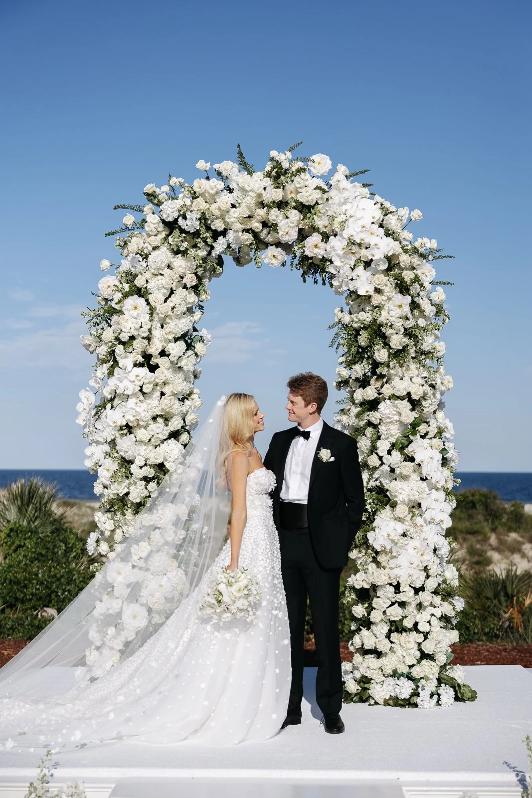 A bride and groom standing under a large arch made of white flowers, facing each other, outdoors by the ocean on a clear day.
