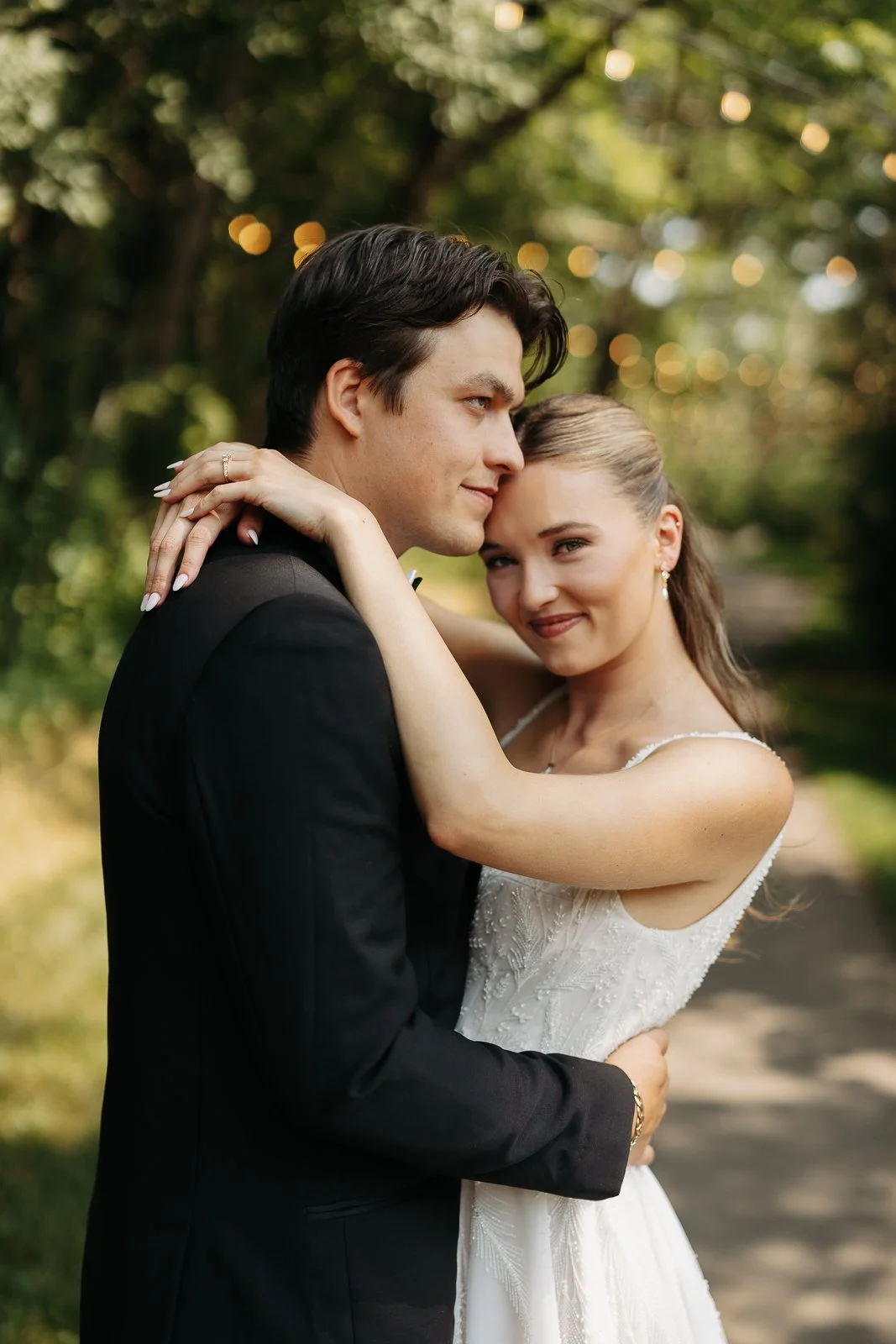 A happy couple embraces outdoors, with the man in a tuxedo and the woman in a wedding dress, surrounded by trees and bokeh lights.