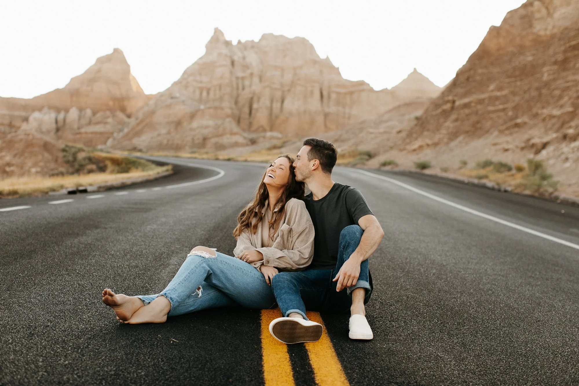 A young couple sitting on an empty desert road with mountains in the background. The woman is smiling with her eyes closed, leaning into the man, who is kissing her on the cheek. They are barefoot and appear happy and relaxed.
