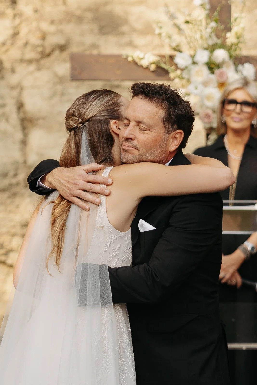 A bride and groom hugging during their wedding ceremony, with a woman in the background.
