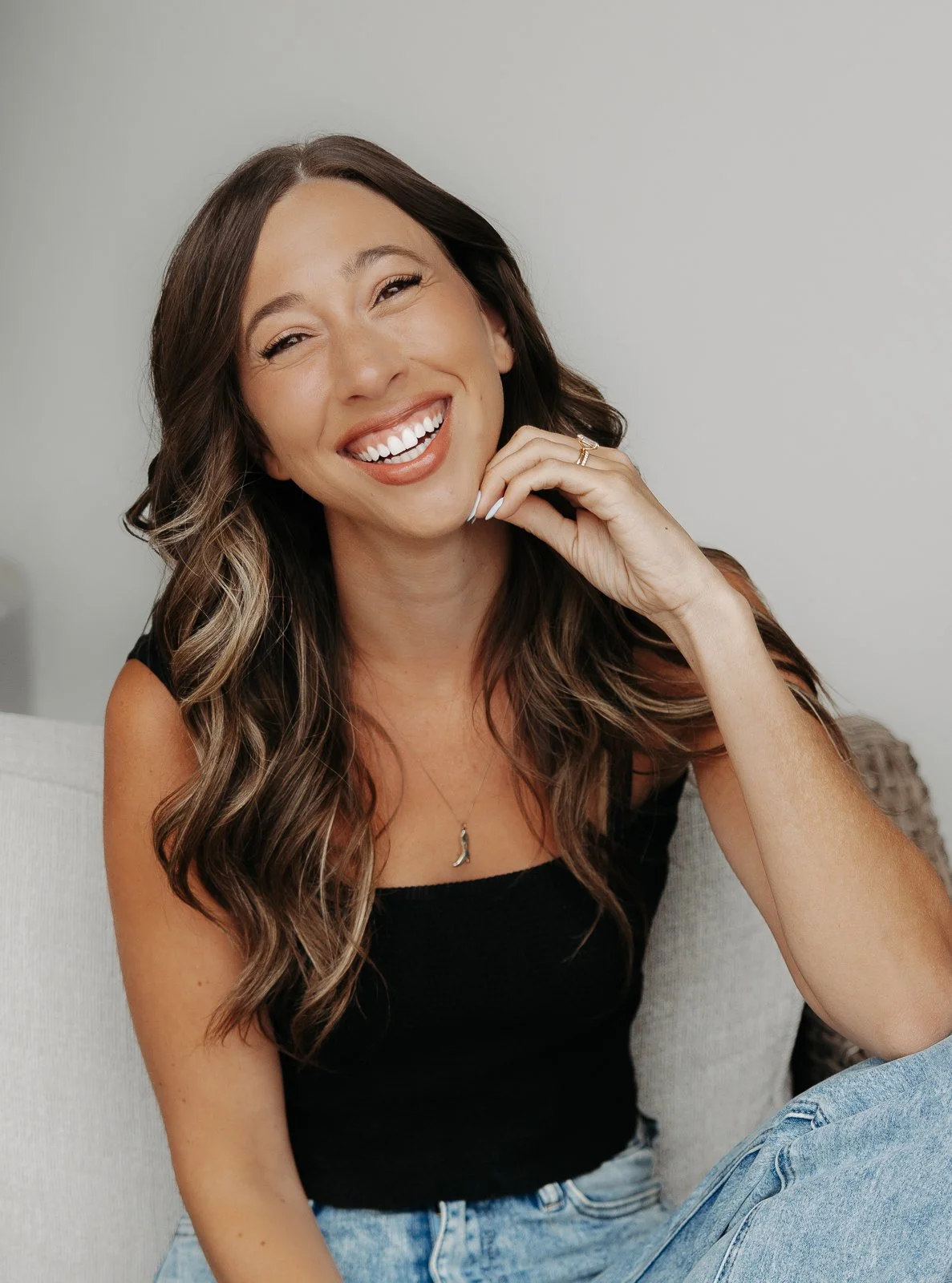 Smiling woman with long wavy hair, wearing a black sleeveless top, gold jewelry, sitting on a couch with a beige pillow in the background.