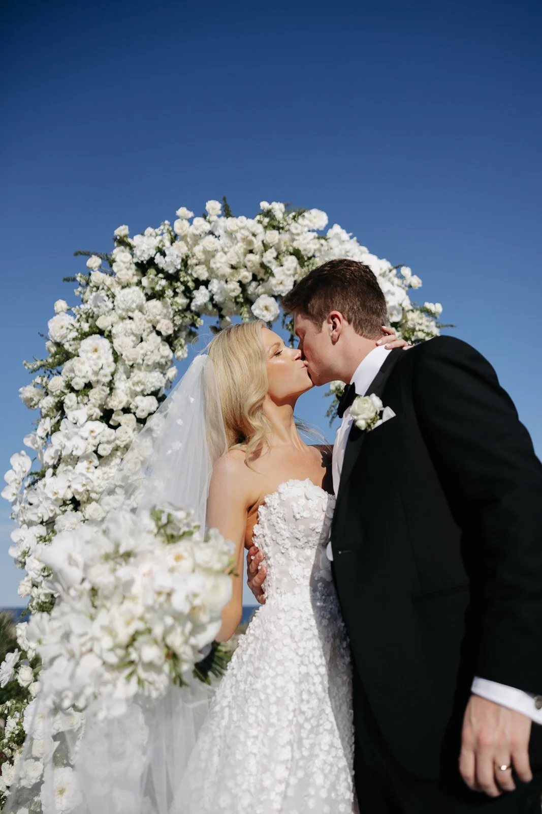 A bride and groom sharing a kiss at their wedding ceremony with a floral arch and blue sky background.