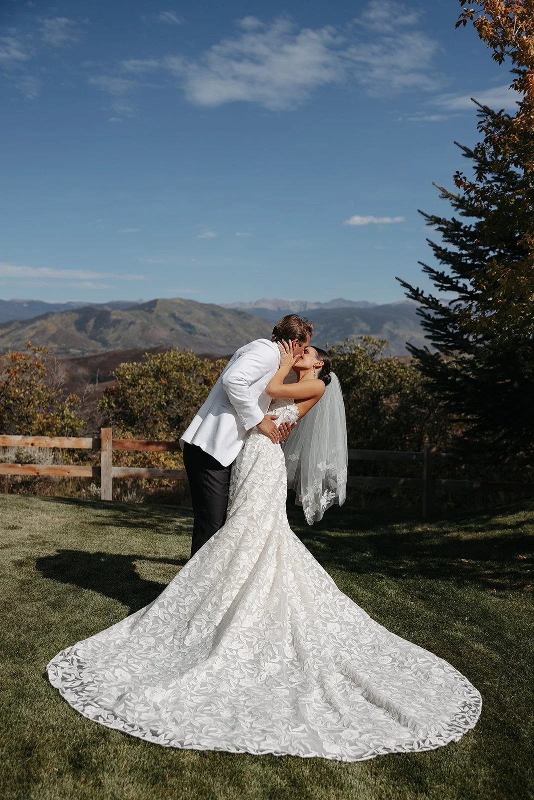 A bride and groom sharing a kiss outdoors on a sunny day, with mountains in the background and trees on the side.