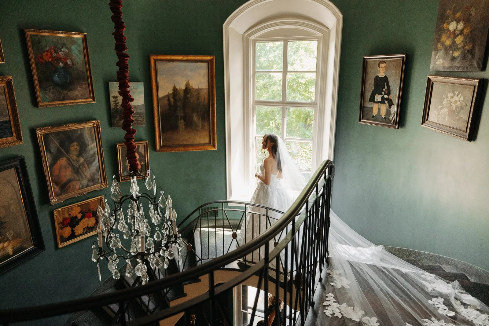 A bride in a white wedding gown and veil standing by a large window in an art-filled gallery or museum, with a staircase and chandelier in the foreground.