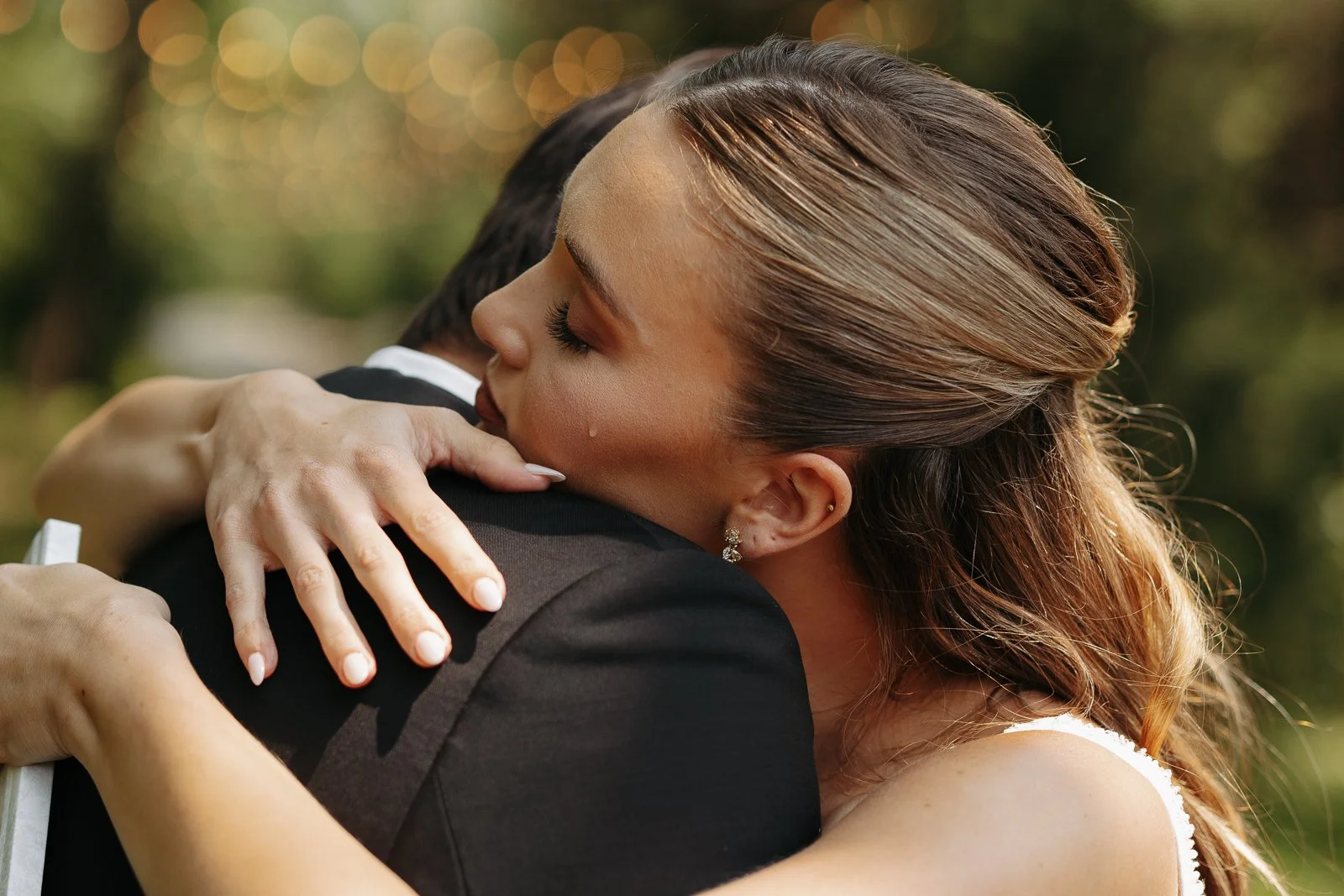 A woman hugging a man, possibly at a wedding or special event, outdoors with blurred greenery and lights in the background.