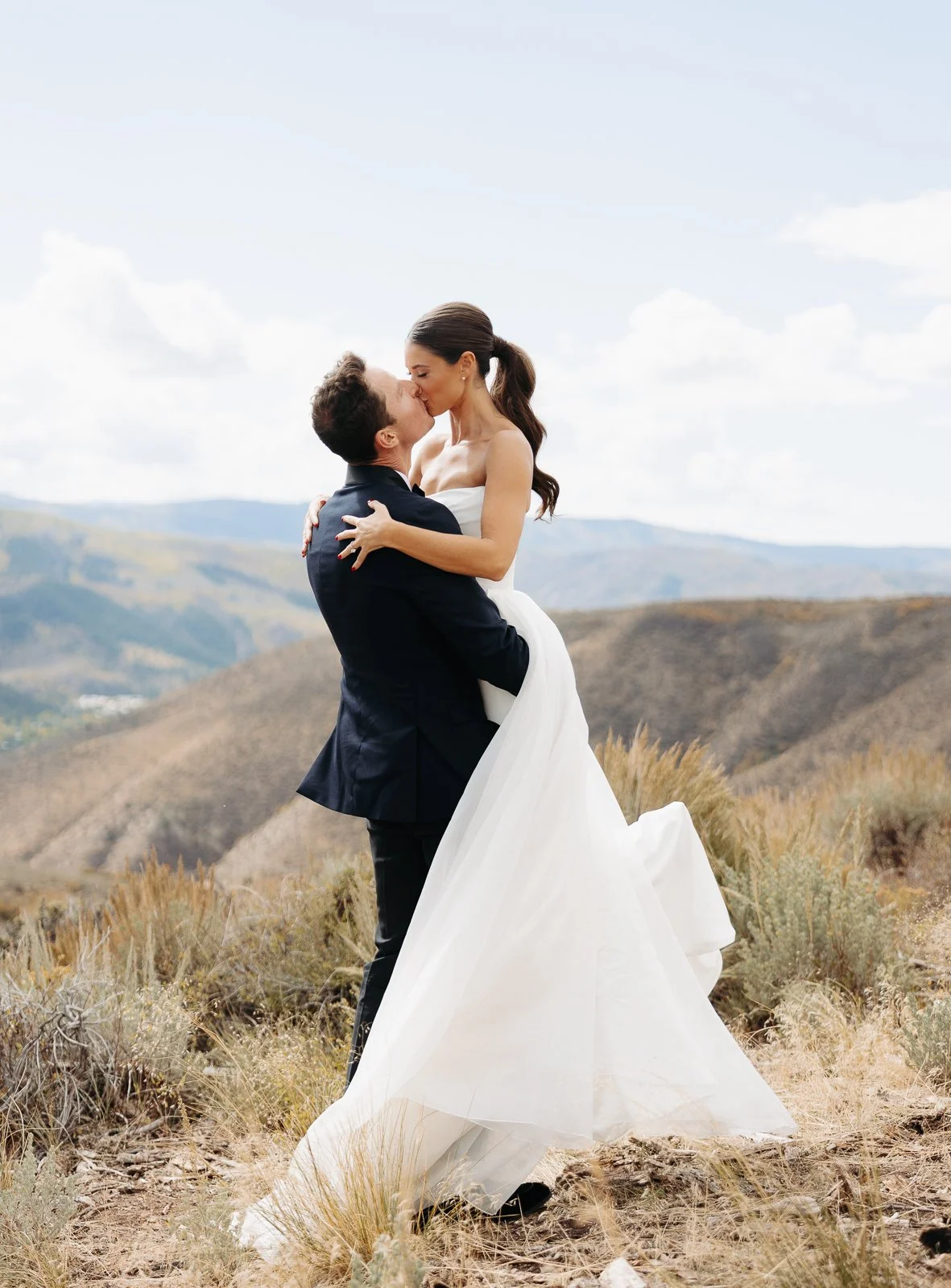 A bride and groom sharing a kiss in a desert landscape with hills in the background, the groom lifting the bride.