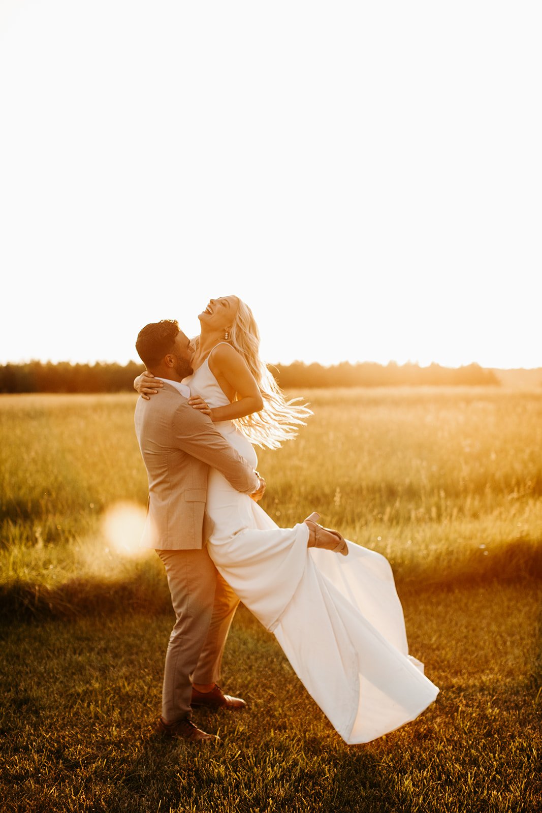 A bride and groom in wedding attire embrace and laugh in a field during sunset, with the groom lifting the bride.