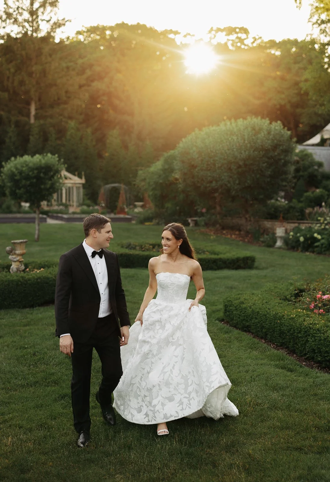 A bride and groom walking on a lush green lawn in a garden during sunset, with the bride wearing a white strapless wedding gown and the groom in a black tuxedo.