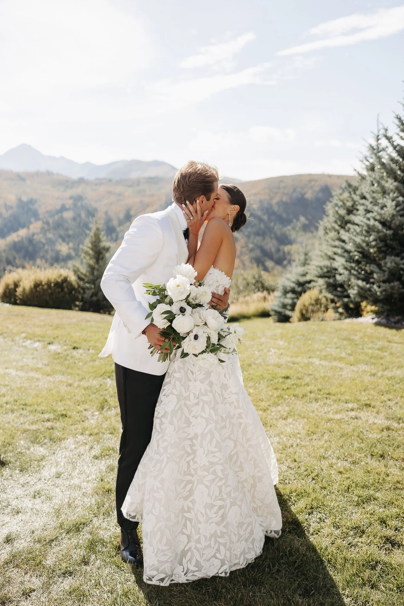 A bride and groom share a kiss outside on a grassy field with mountains and trees in the background. The bride is wearing a strapless white wedding gown and holding a bouquet of white flowers, while the groom is dressed in a white tuxedo jacket and b