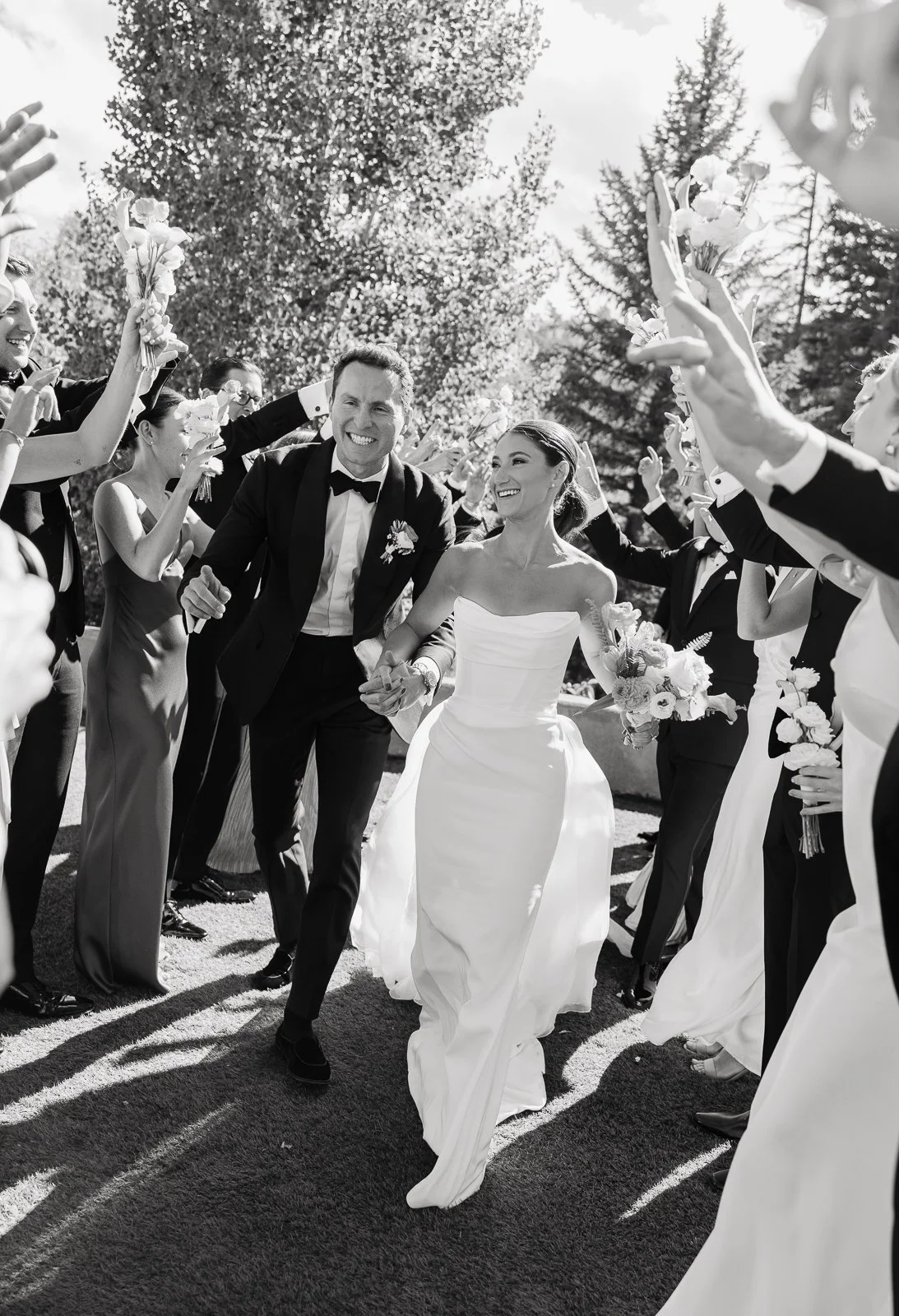 A black and white photo of a bride and groom walking hand in hand through a line of people celebrating at their wedding outdoors, with trees in the background.