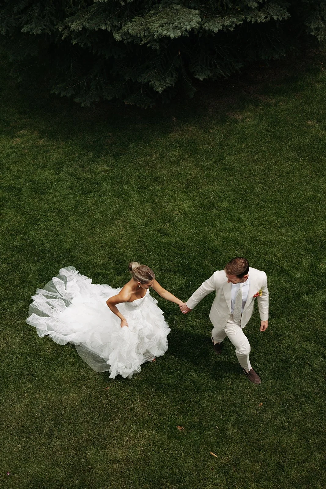 A bride and groom holding hands on a grassy lawn, with the bride wearing a white wedding dress and the groom in a white suit.