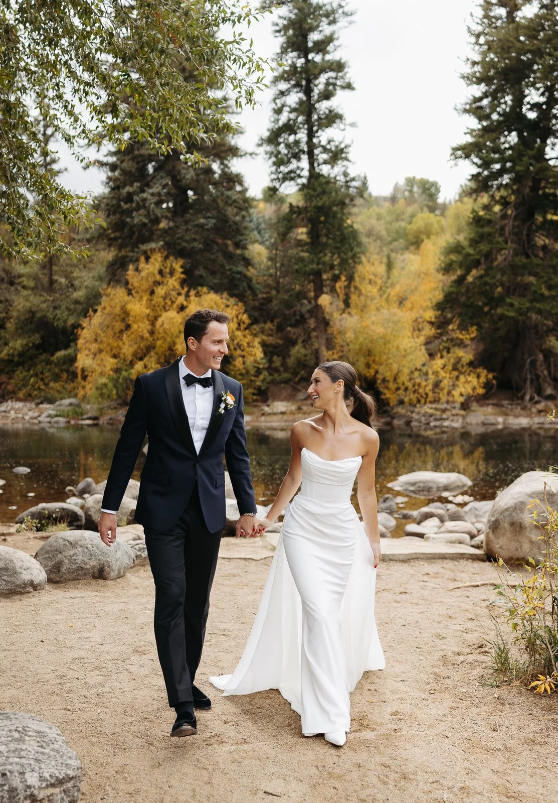 A bride and groom walking hand in hand along a sandy shore by a lake, surrounded by trees with autumn foliage, smiling and looking at each other.
